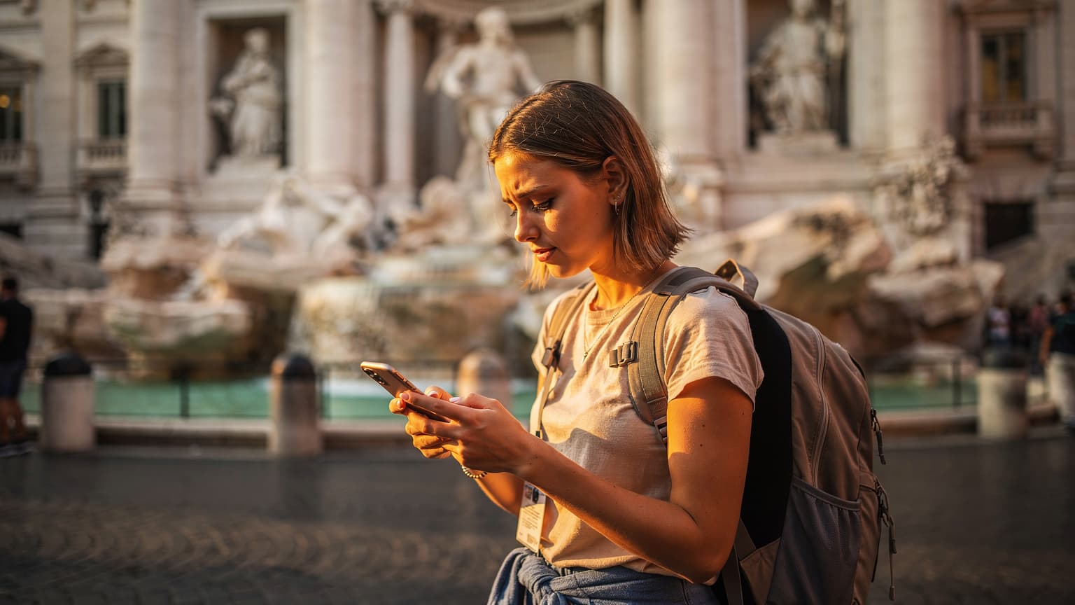 Una viajera activa revisa su teléfono en la plaza de la Fontana di Trevi en Roma, con la fuente y la arquitectura histórica de fondo.