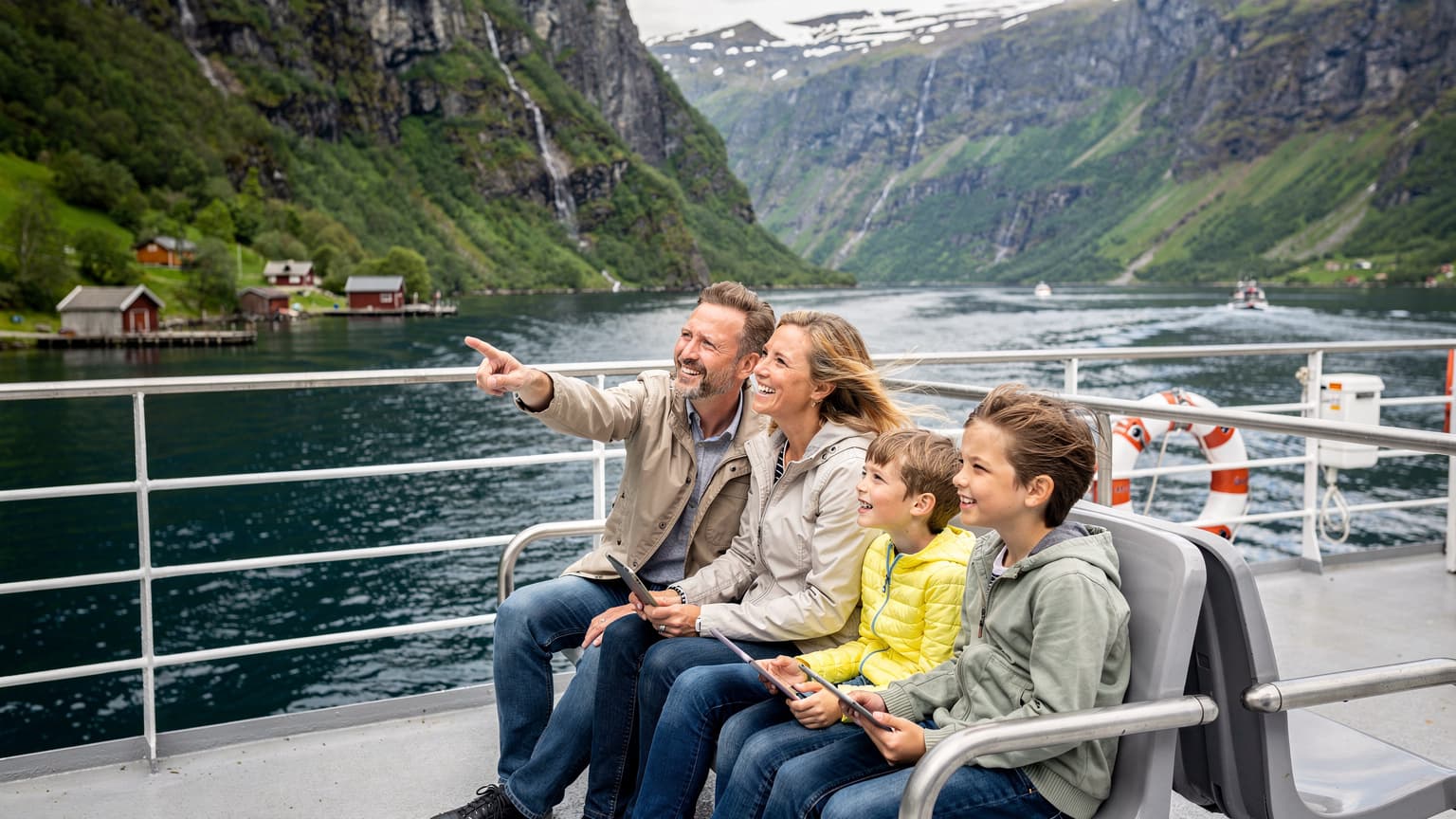 En familie på fire, to voksne og to barn (4 og 10 år), sitter på en ferge som krysser en norsk fjord. De ser ut over det majestetiske landskapet med bratte fjellsider og små hus langs vannkanten. Barna holder hver sin iPad og smiler, mens foreldrene peker på noe i det fjerne. Været er lyst og klart, og solen skinner.