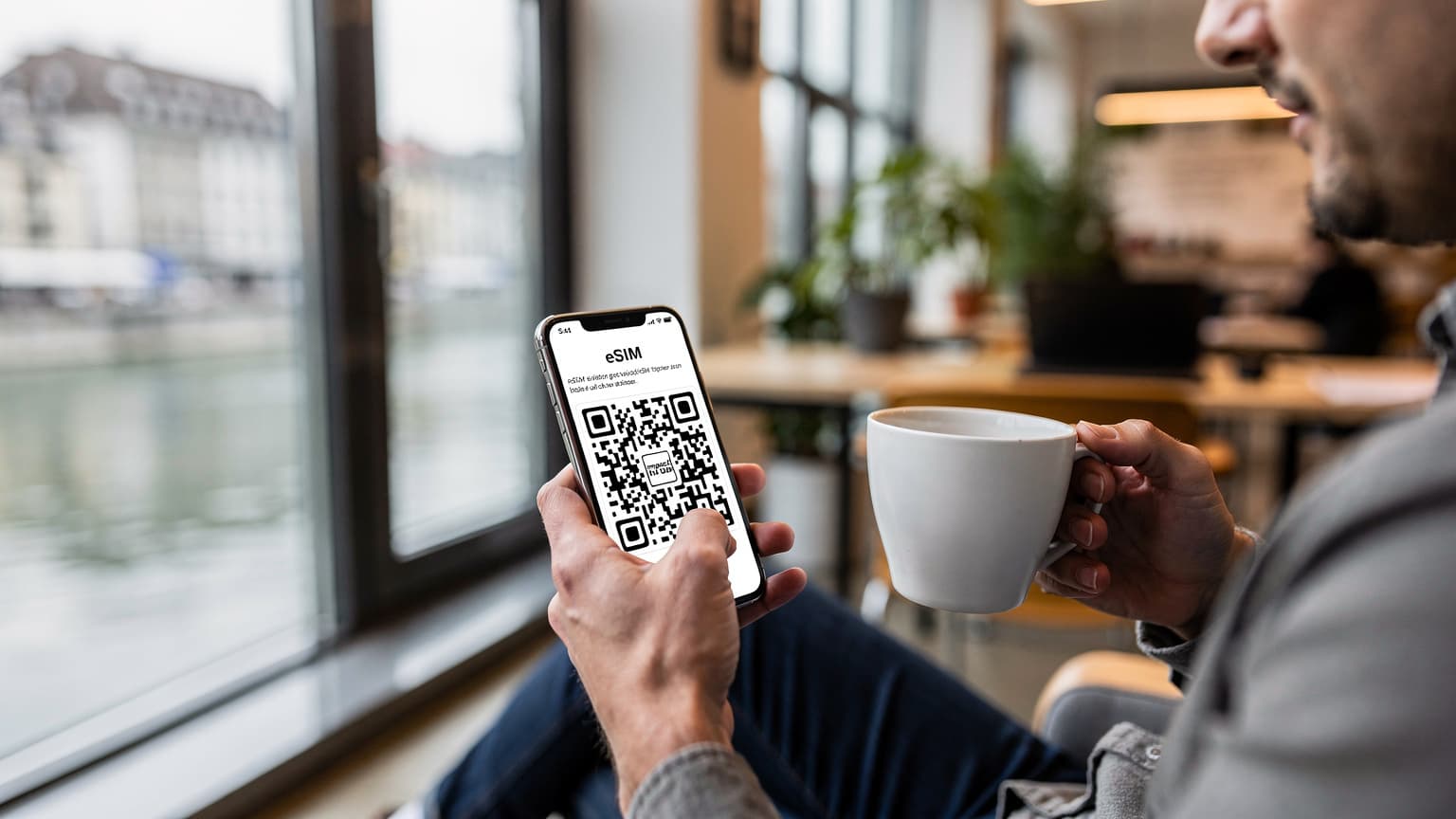 Un viajero nómada digital configurando su eSIM en un smartphone mientras toma un café en un coworking moderno en Zúrich, con vistas a la ciudad.