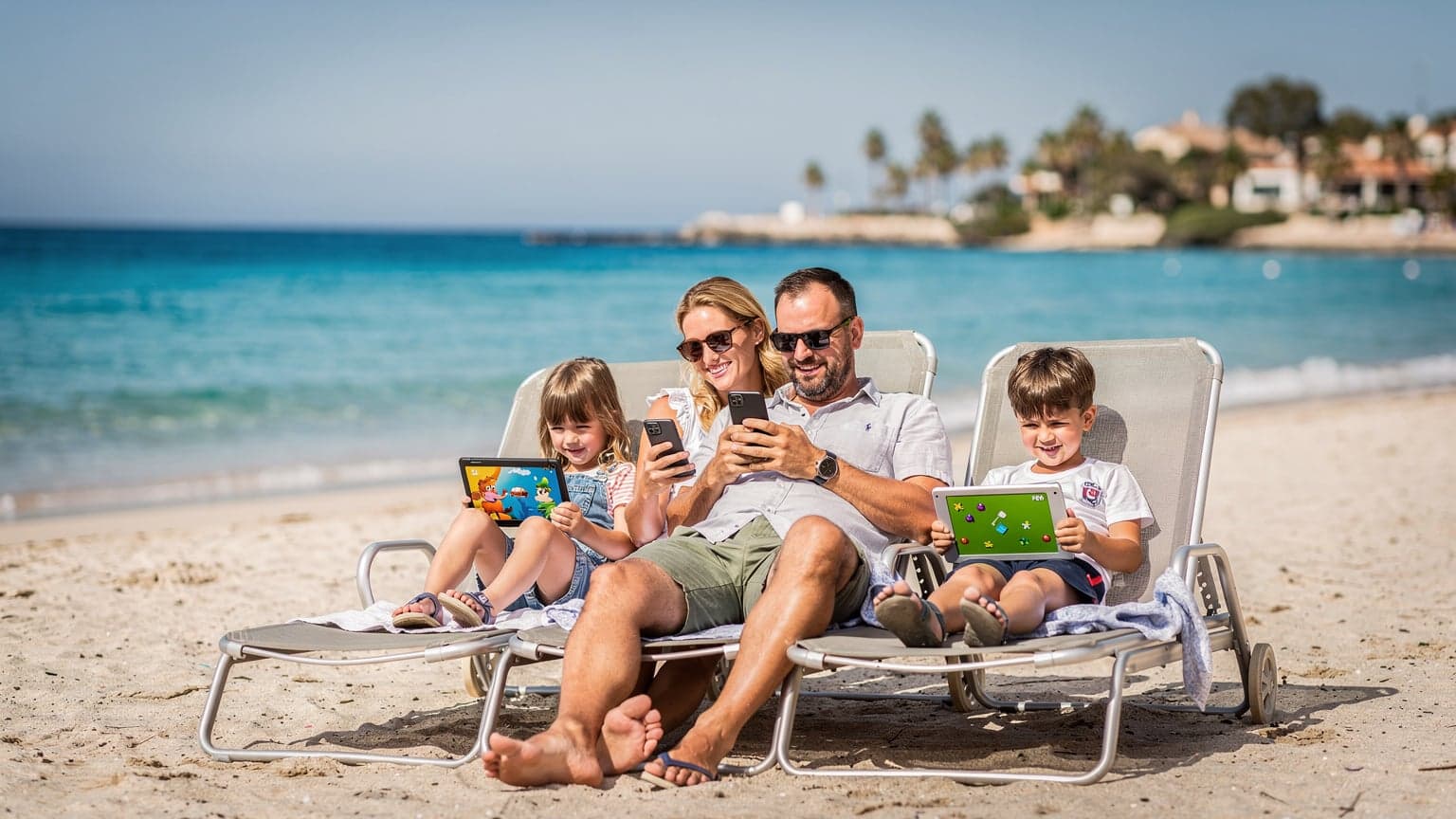 Rodzina z dwójką dzieci, trzymająca smartfony i tablety, relaksująca się na leżakach na słonecznej plaży na Majorce, z błękitnym morzem i palmami w tle.