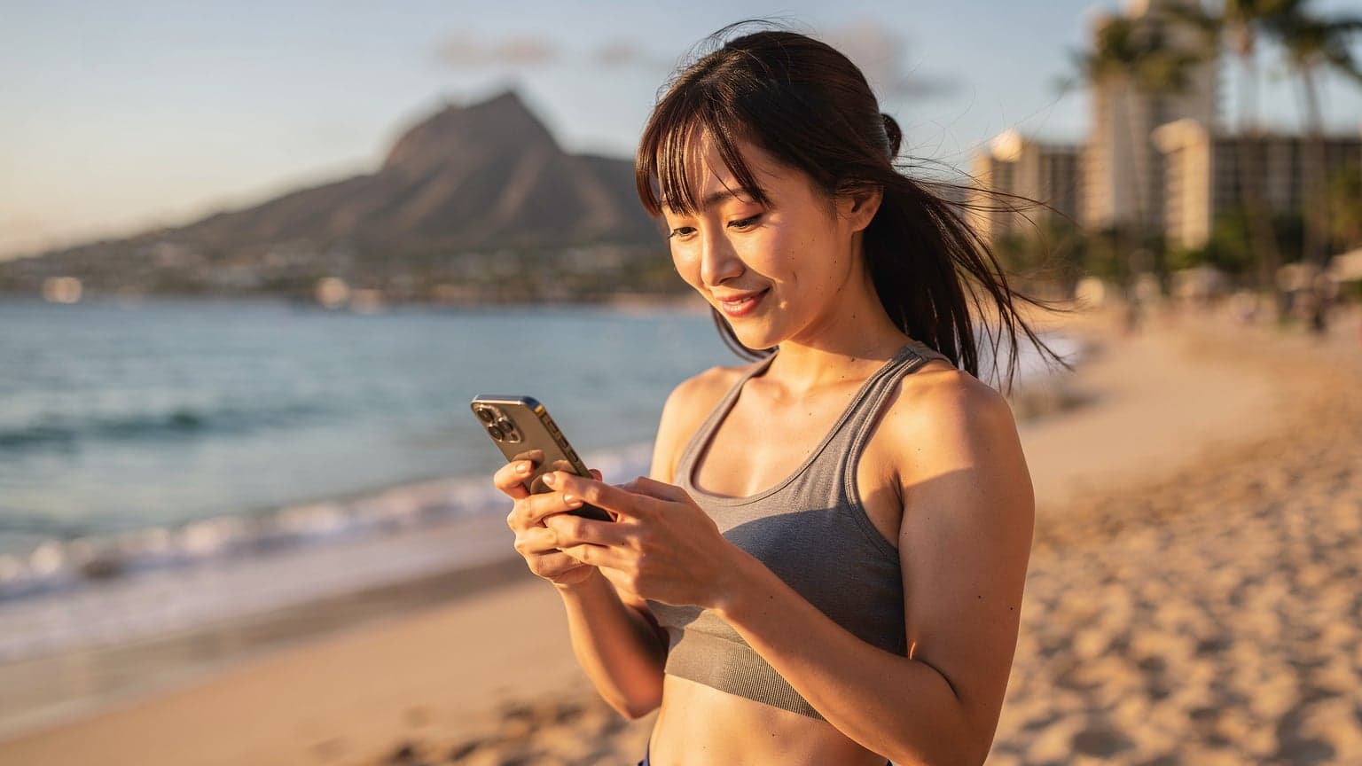 夏休みの旅行者が、ハワイのワイキキビーチを背景にスマートフォンでeSIMの設定を確認している様子。家族や友人との連絡をスムーズにするために、ネットワーク接続を最適化している。