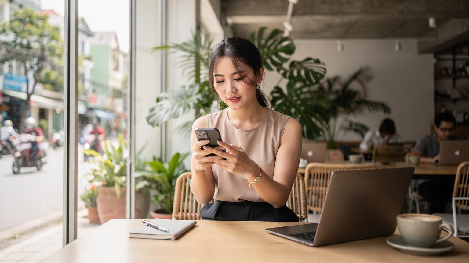 Một phụ nữ trẻ đang ngồi trong quán cà phê hiện đại ở quận Thảo Điền, Thành phố Hồ Chí Minh, với máy tính xách tay và điện thoại thông minh trên bàn, kiểm tra eSIM trước chuyến đi.