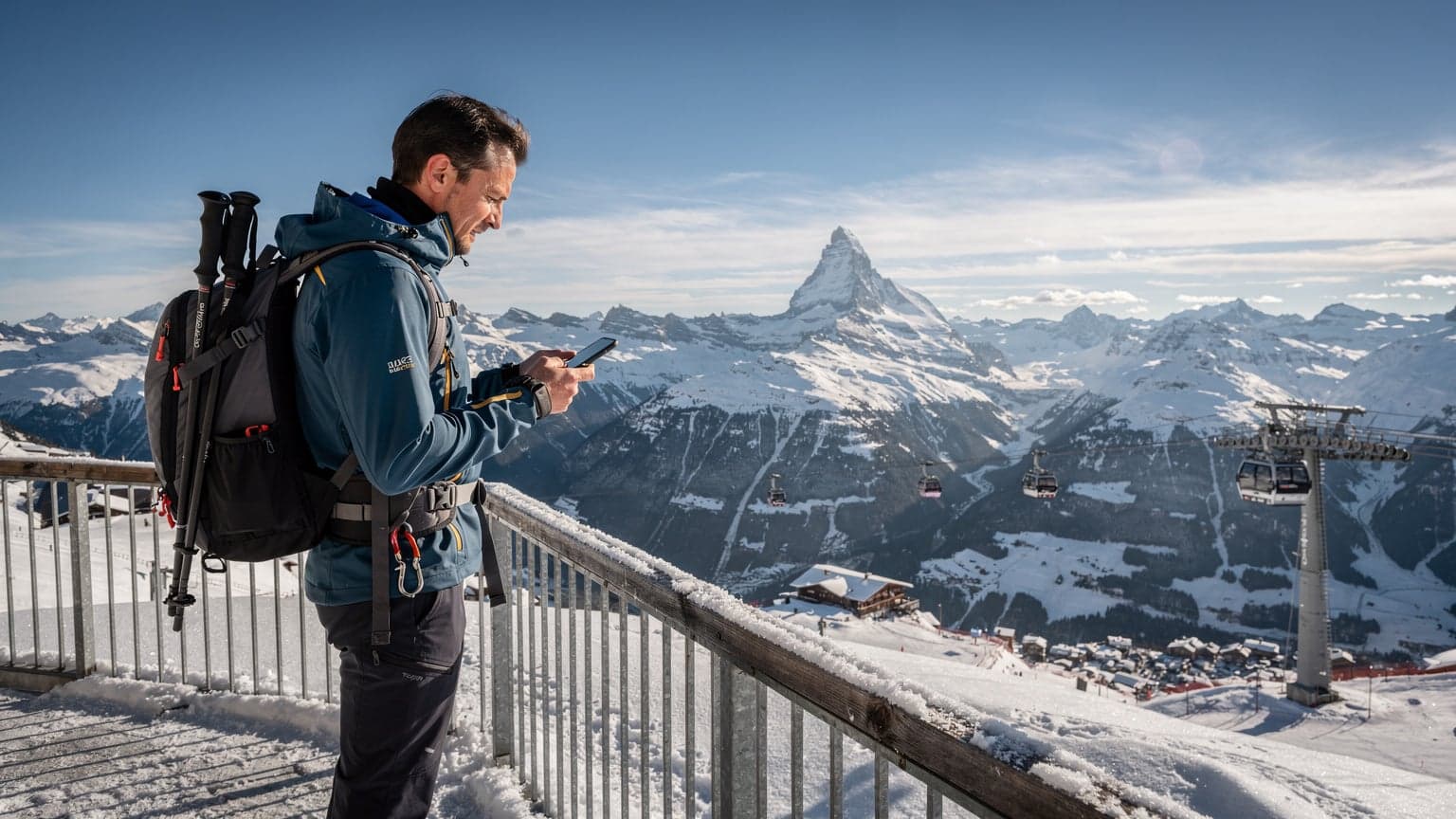 Una persona con mochila de senderismo y bastones de esquí mirando un mapa digital en su teléfono, con los Alpes suizos nevados de fondo y un cielo azul claro.