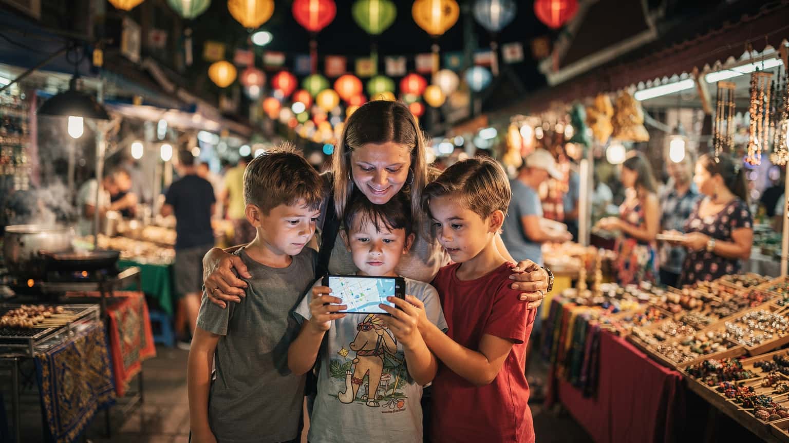 Une mère souriante et ses deux enfants, âgés d'environ 8 et 10 ans, naviguent dans un marché de nuit animé à Chiang Mai, Thaïlande, tous regardant attentivement un smartphone affichant une carte. Le marché est éclairé par des lanternes colorées et des néons, avec des étals de nourriture et d'artisanat en arrière-plan.