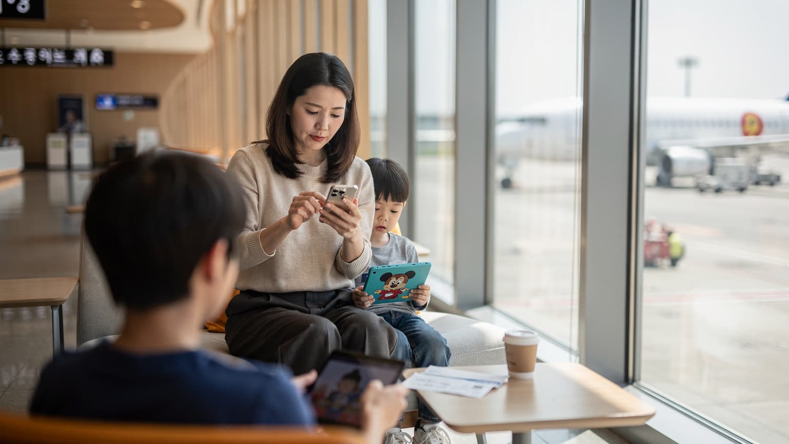 서울의 김포공항 출국장 라운지에서 한 어머니가 아이와 함께 태블릿으로 애니메이션을 보며 스마트폰으로 Cellesim eSIM을 활성화하는 모습.