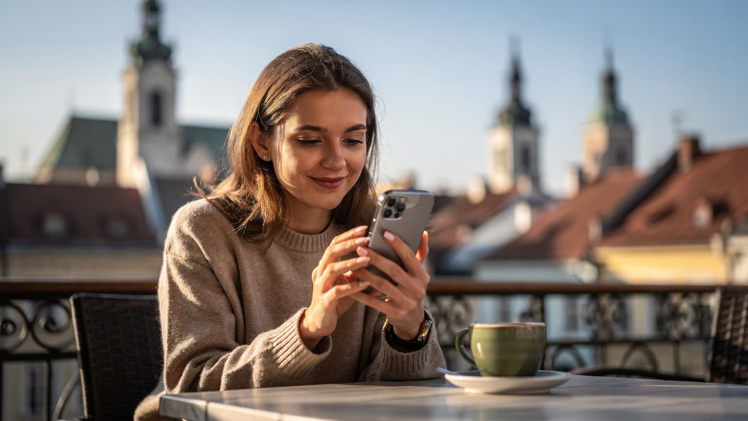 Жінка з iPhone 17 Pro Max у руках, посміхається, сидячи на терасі кафе у Львові, на задньому плані видно старовинні дахи міста.