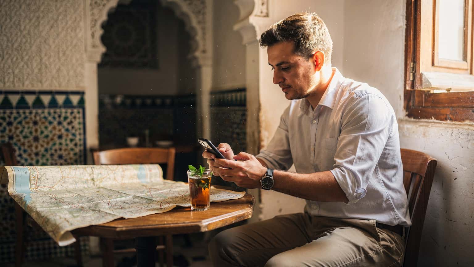 A person holding a smartphone and looking at a historical map while sitting at a traditional cafe in the Casbah of Algiers, surrounded by detailed Arabic architecture.