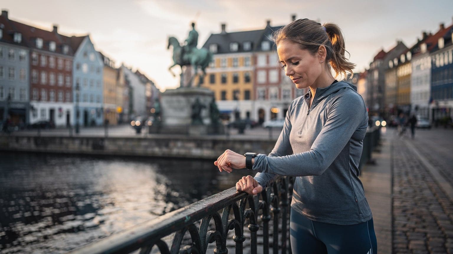En kvinde i løbetøj tjekker sit Apple Watch for notifikationer, mens hun står på en bro i København med Højbro Plads i baggrunden, badet i blødt morgenlys.