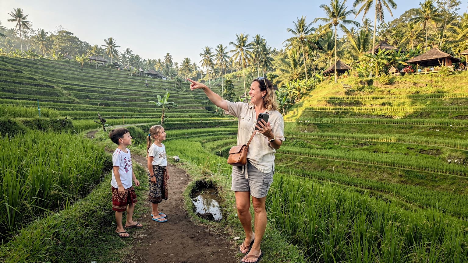 Seorang ibu tersenyum sambil memegang ponselnya, menunjuk ke arah pemandangan sawah terasering di Ubud, Bali, dengan anak-anaknya bermain di latar belakang yang cerah.