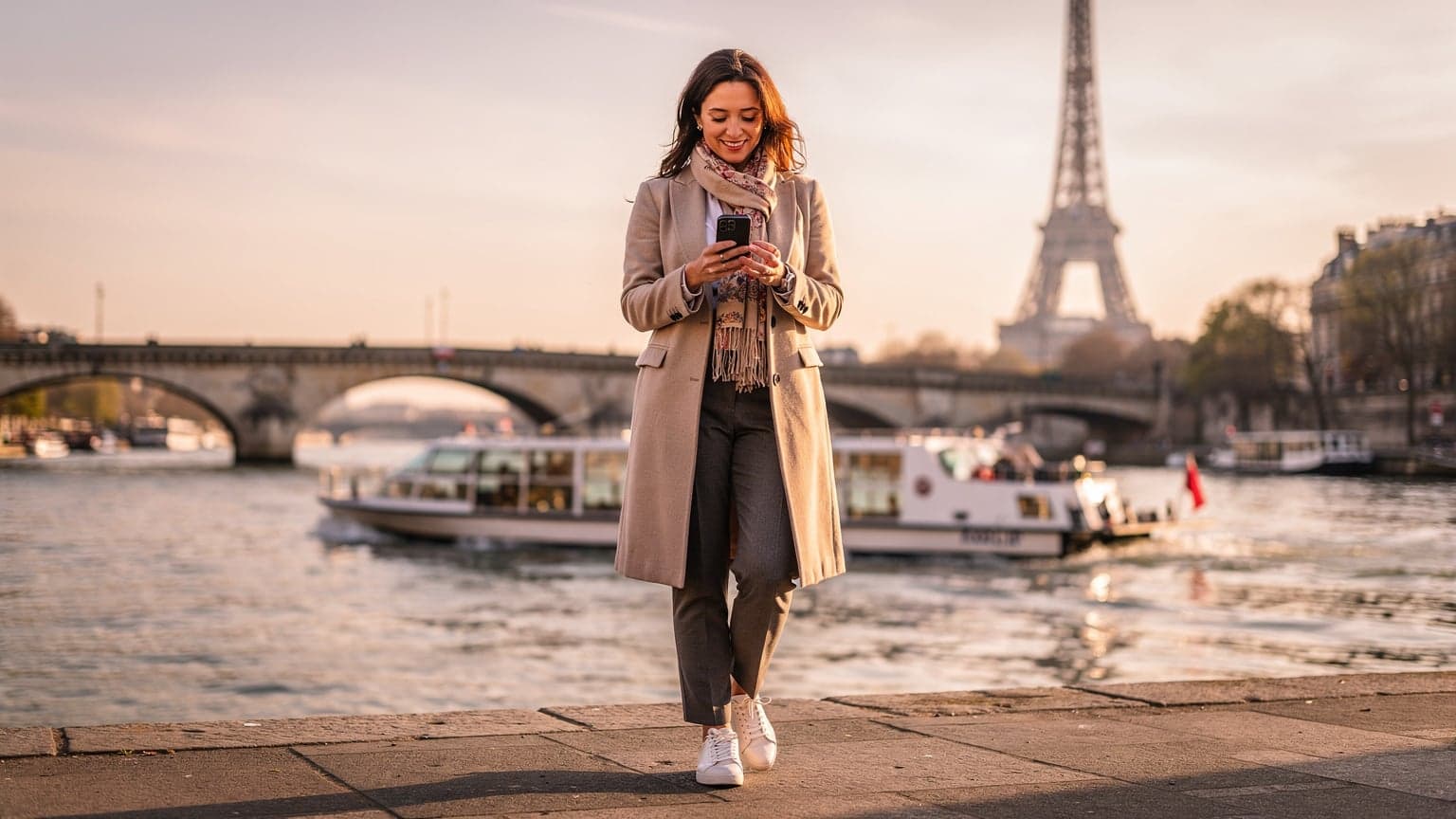 Une femme souriante consulte son téléphone sur un pont surplombant la Seine, avec la Tour Eiffel en arrière-plan et un bateau-mouche passant en dessous, au lever du soleil.