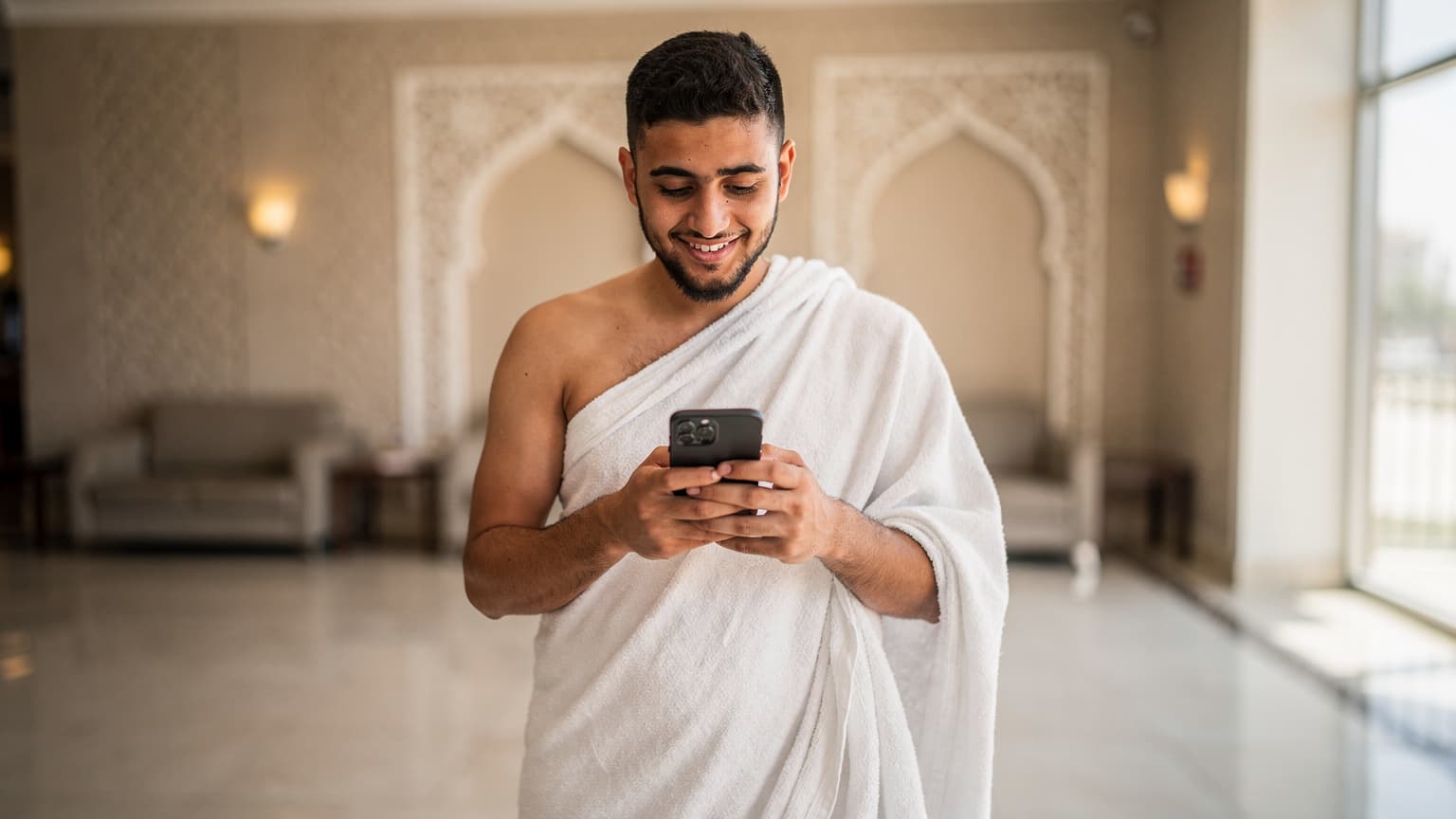 معتمر مصري شاب, يرتدي ملابس الإحرام البيضاء, يستخدم هاتفه الذكي داخل فندق بسيط في مكة المكرمة, يبحث عن اتجاهات أو يتواصل مع عائلته بابتسامة.