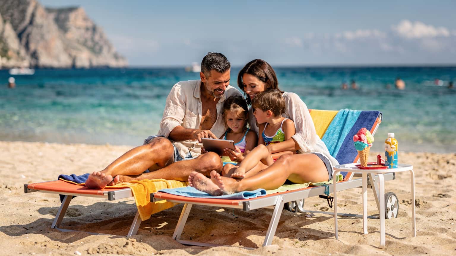 Una famiglia italiana, composta da due genitori e due bambini di circa 8 e 10 anni, sorride mentre guarda un tablet su una sdraio in una spiaggia assolata in Sicilia, con il mare azzurro sullo sfondo e un gelato mezzo sciolto sul tavolino vicino.