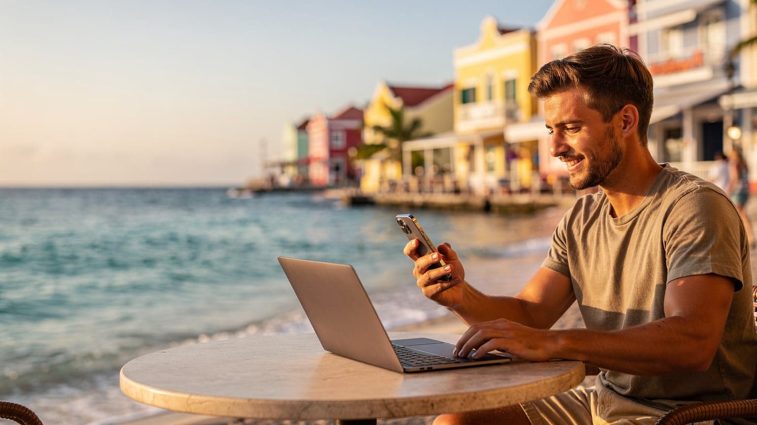 Een vrouw met een zonnehoed kijkt naar haar telefoon op een terras met uitzicht op het kristalheldere water en kleurrijke gebouwen van Willemstad, Curaçao.