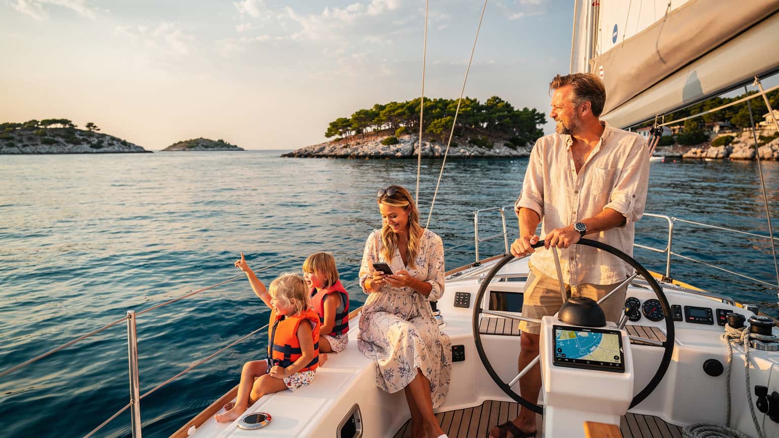 En familj med två barn sitter på däck på en segelbåt i Kroatiska skärgården, med kristallklart vatten och gröna öar i bakgrunden, solen skiner och barnen tittar på en iPad.