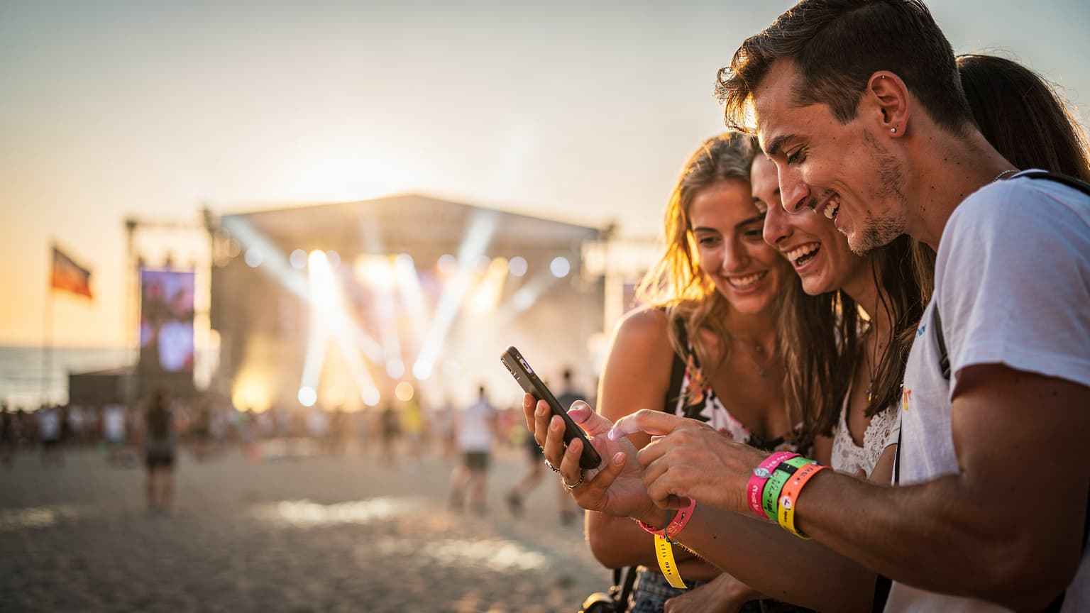 O tânără zâmbitoare, cu un telefon în mână, verificând programul festivalului la Neversea, cu scena principală luminoasă în fundal, la apus.
