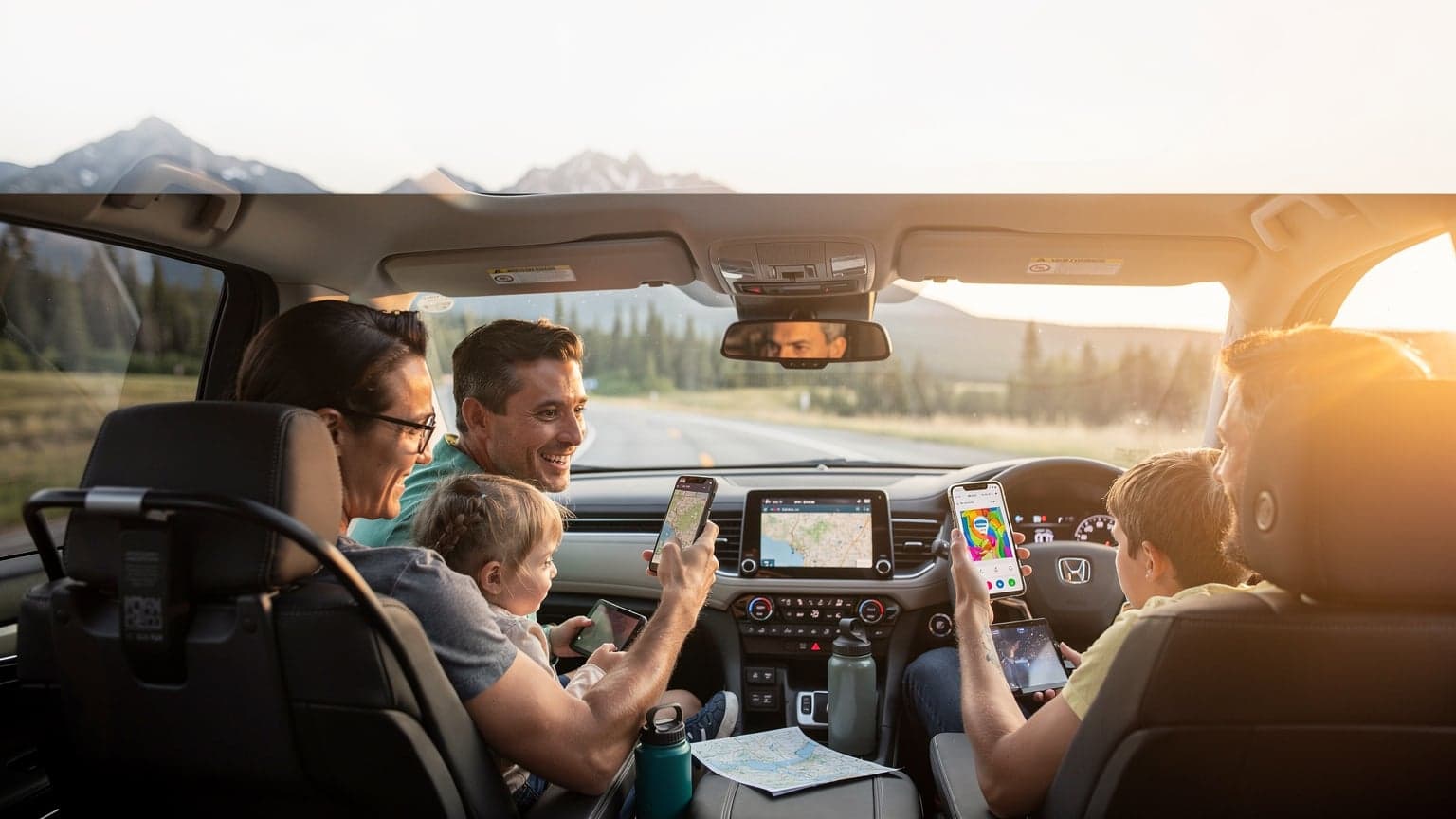 A family laughing in a car, with a child in the back seat holding a tablet, driving through a scenic North American mountain pass.