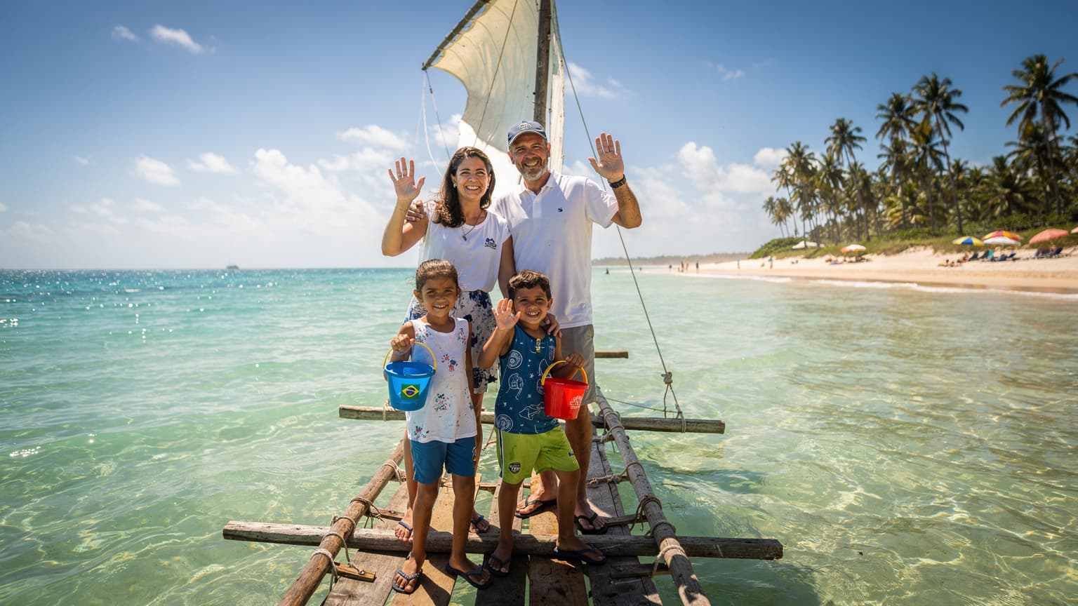 Uma família, pai, mãe e duas crianças, sorrindo e acenando de uma jangada colorida na praia de Porto de Galinhas, Pernambuco, com águas claras e coqueiros ao fundo, sob um céu azul vibrante.