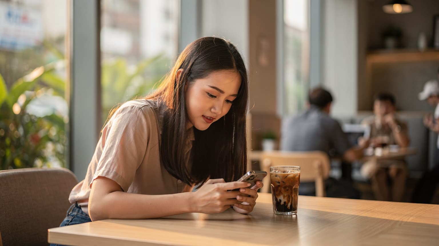 Một phụ nữ Việt Nam trẻ tuổi ngồi tại bàn trong một quán cà phê hiện đại ở Sài Gòn, tập trung vào điện thoại di động của mình với một ly cà phê đá bên cạnh.