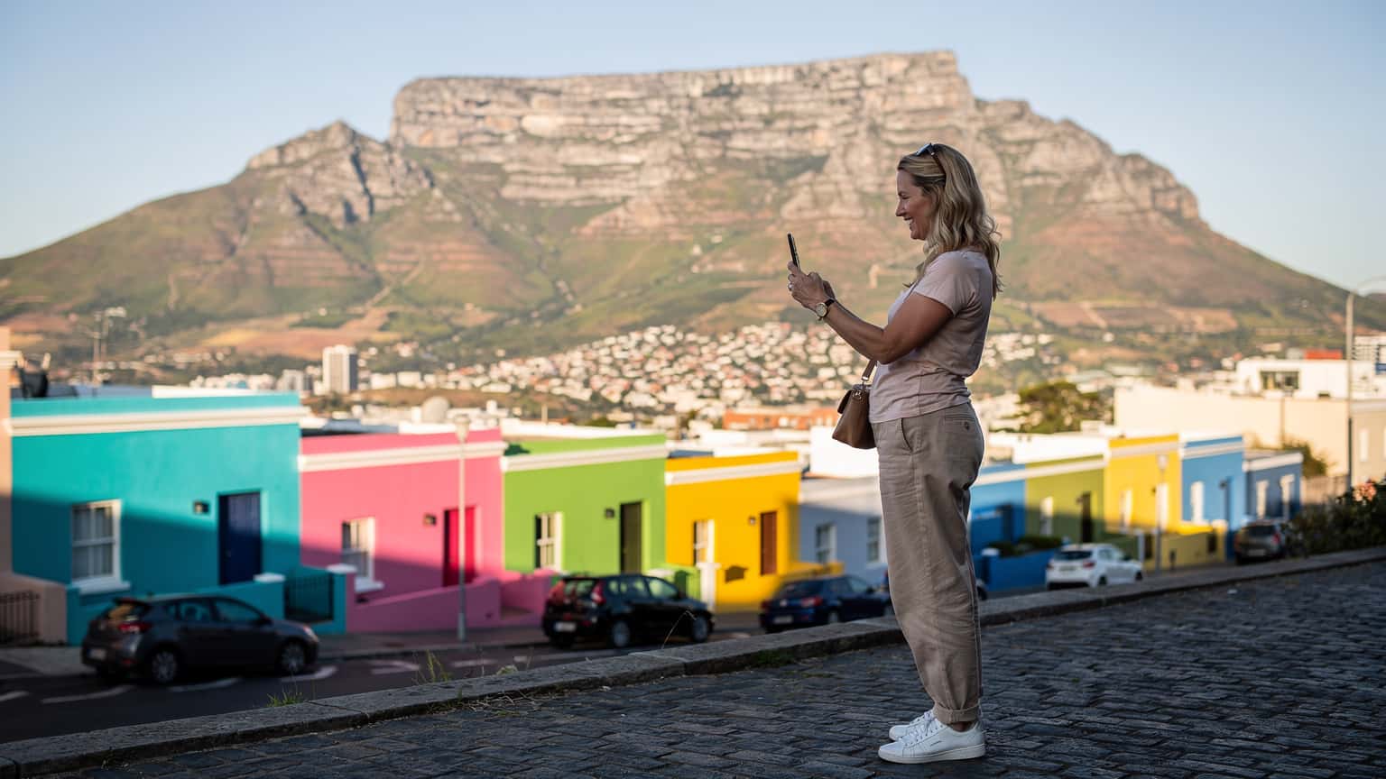 Een vrouw met een smartphone in haar hand, lachend terwijl ze een foto maakt van de Tafelberg in Kaapstad, met de stad beneden en een helderblauwe lucht erboven.