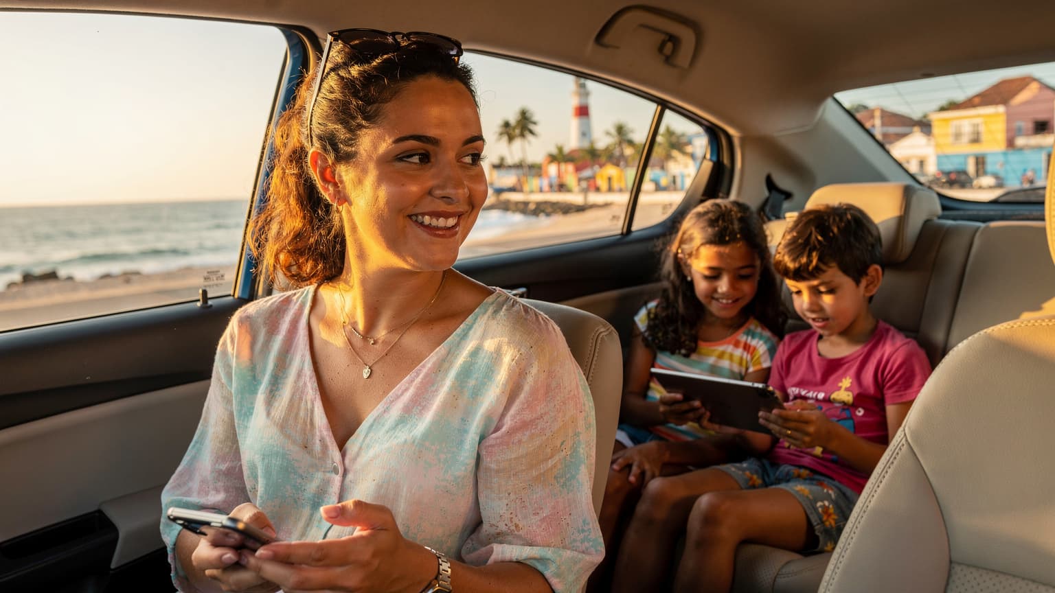 Uma mãe brasileira sorridente, com óculos de sol na cabeça, segura um smartphone enquanto seu filho e filha pequenos assistem a algo em um tablet no banco de trás de um carro alugado, com uma paisagem costeira vibrante de Salvador, Bahia, ao fundo, sob a luz do sol da tarde.