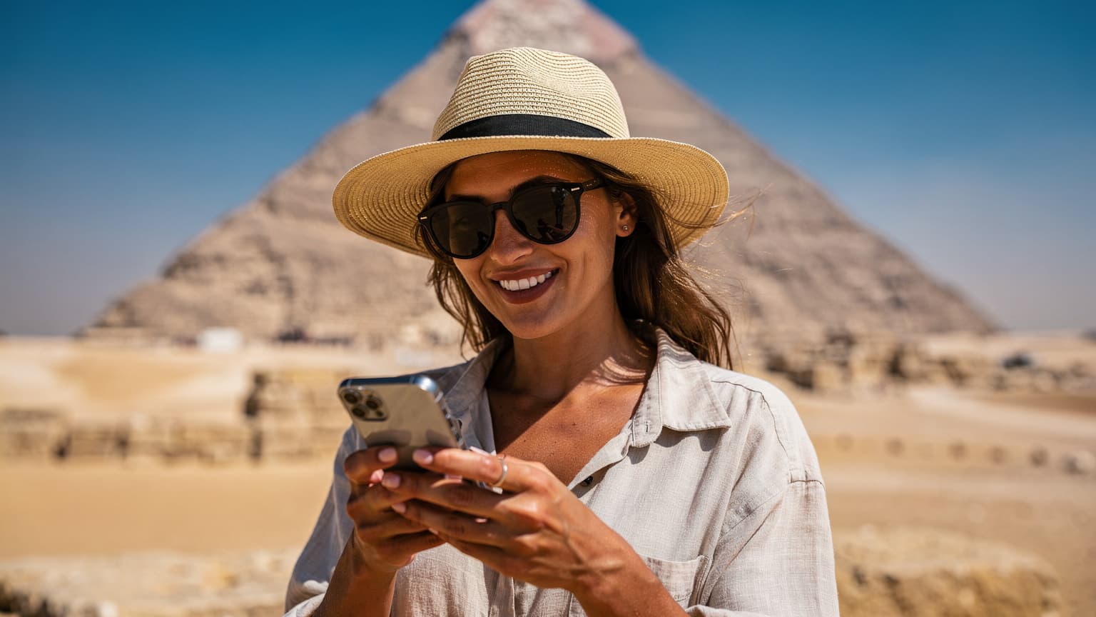 Une femme souriante, d'origine française, consulte son téléphone portable à l'extérieur, avec les pyramides de Gizeh en arrière-plan, sous un ciel bleu clair. Elle porte un chapeau de soleil et des lunettes de soleil, et l'architecture antique est visible mais légèrement floue derrière elle.