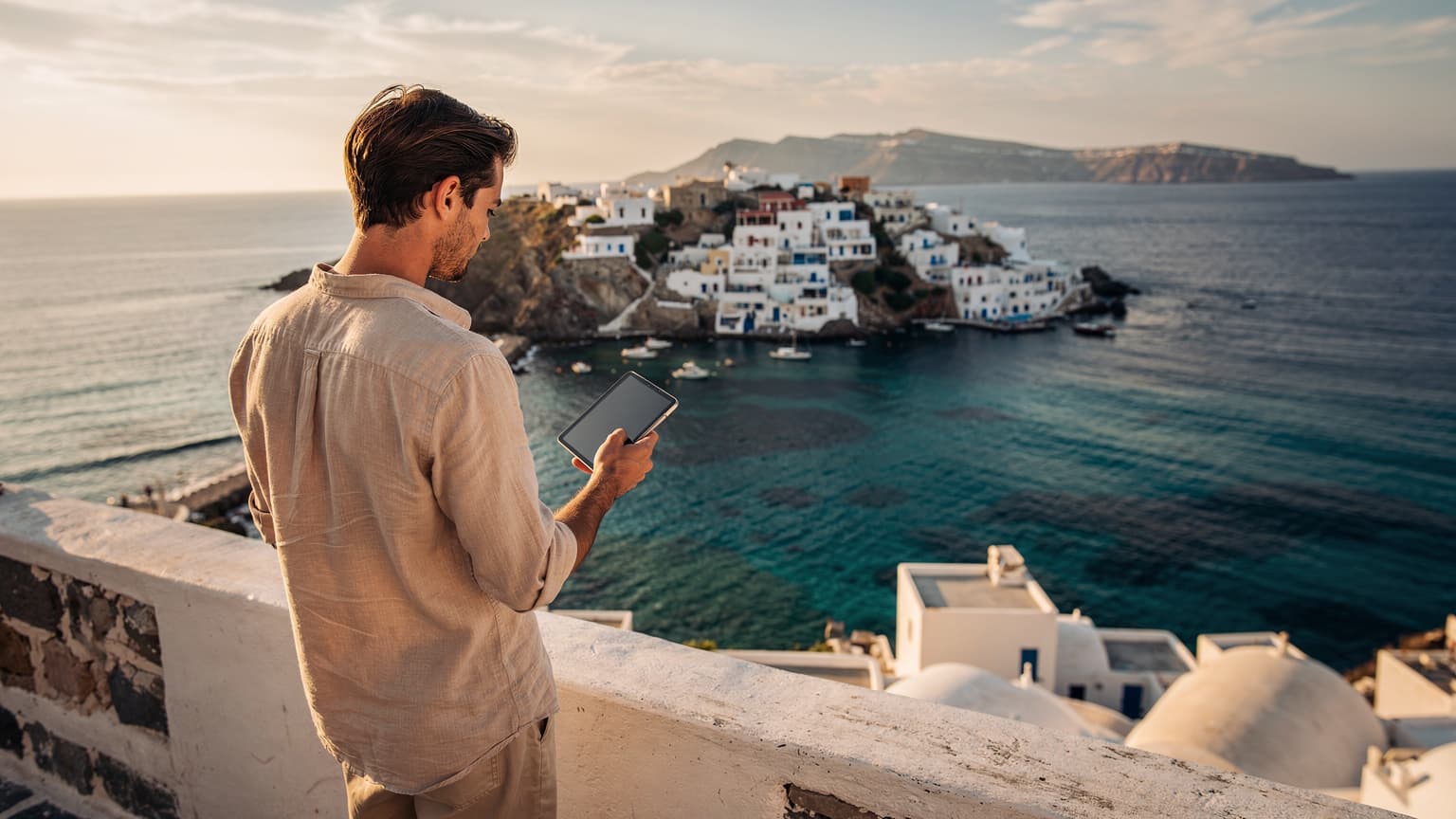 A person on a sunny Mediterranean terrace looking at a smartphone, with turquoise water and an island village in the background, symbolizing connected travel.