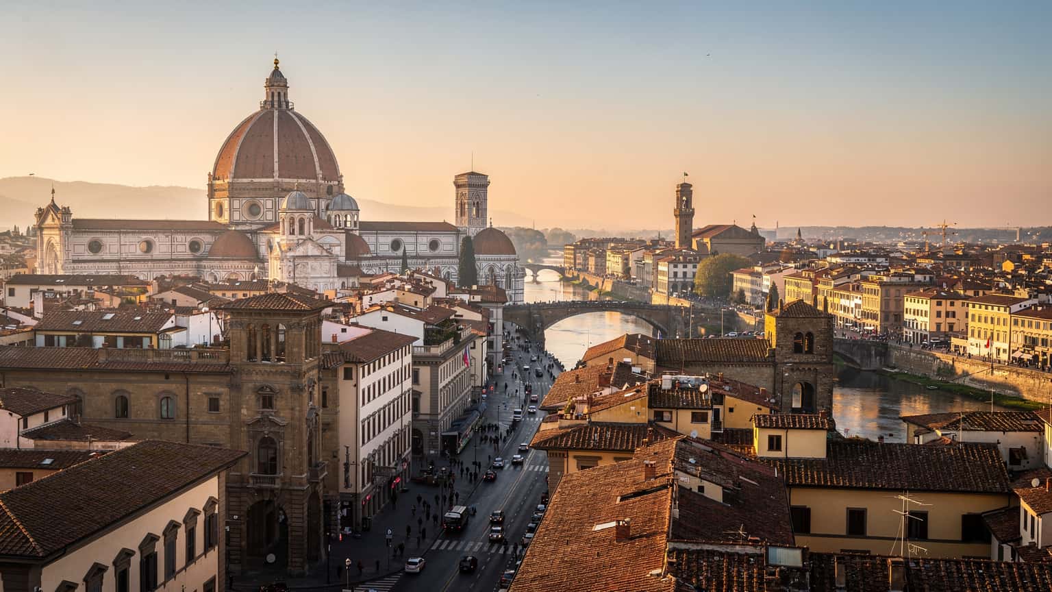 A wide, cinematic shot of an iconic Italian city, like Florence or Rome, at golden hour. Historic buildings are illuminated by warm, natural light, conveying a sense of travel and discovery.