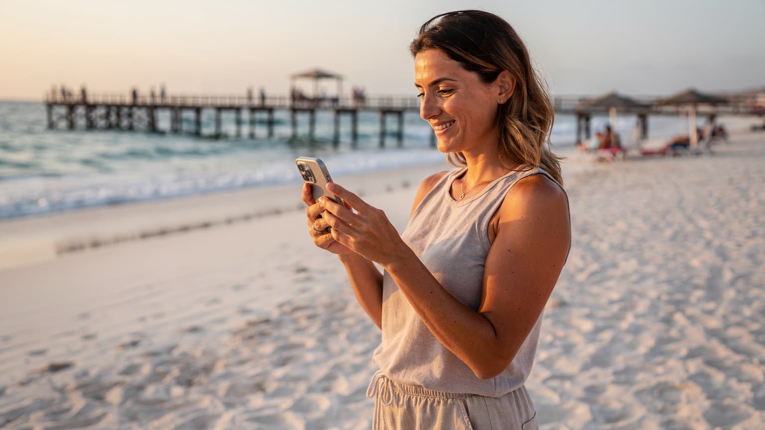 Uma mulher sorridente em uma praia de areia branca na Ilha do Sal, Cabo Verde, olhando para o seu iPhone enquanto o sol da tarde se põe no horizonte.
