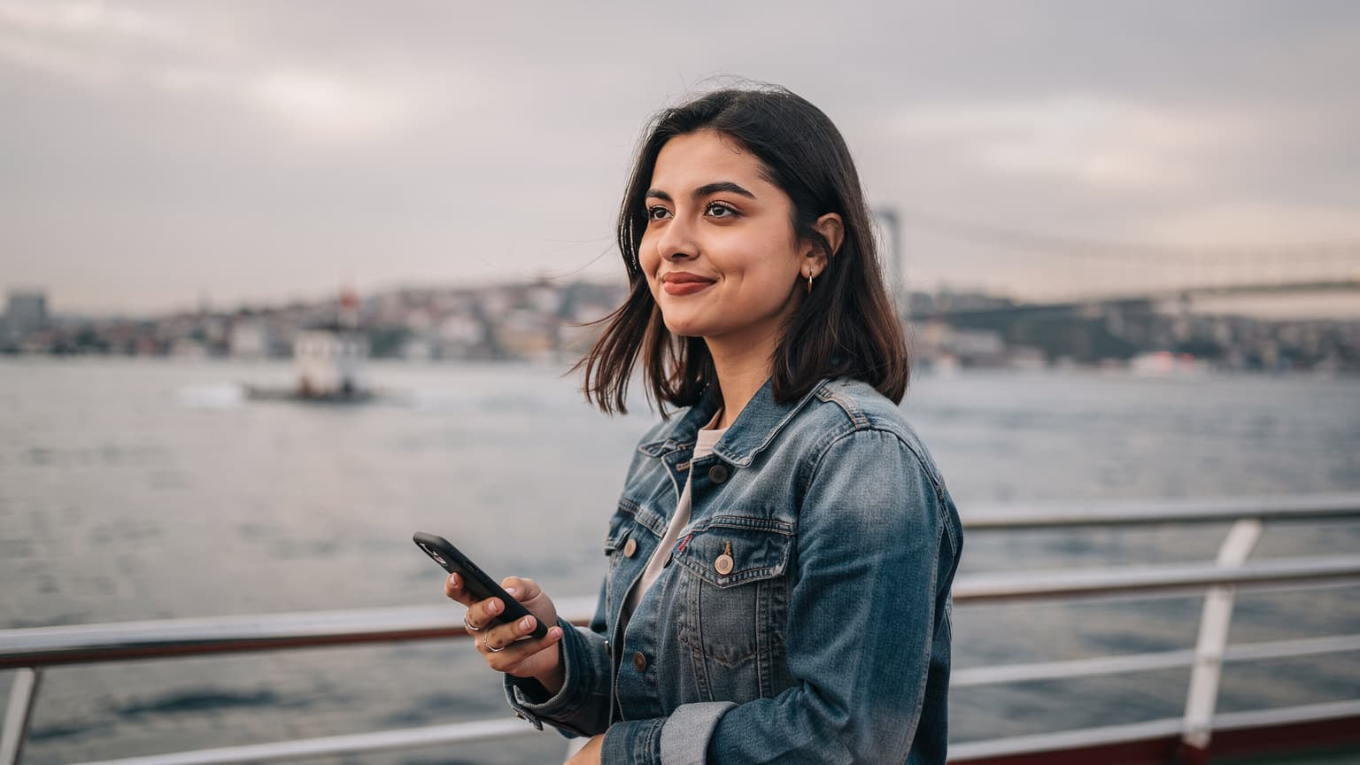 İstanbul'da bir vapur güvertesinde, elinde akıllı telefon tutan bir kadın, şehrin simgesel yapılarına bakarken gülümsüyor.