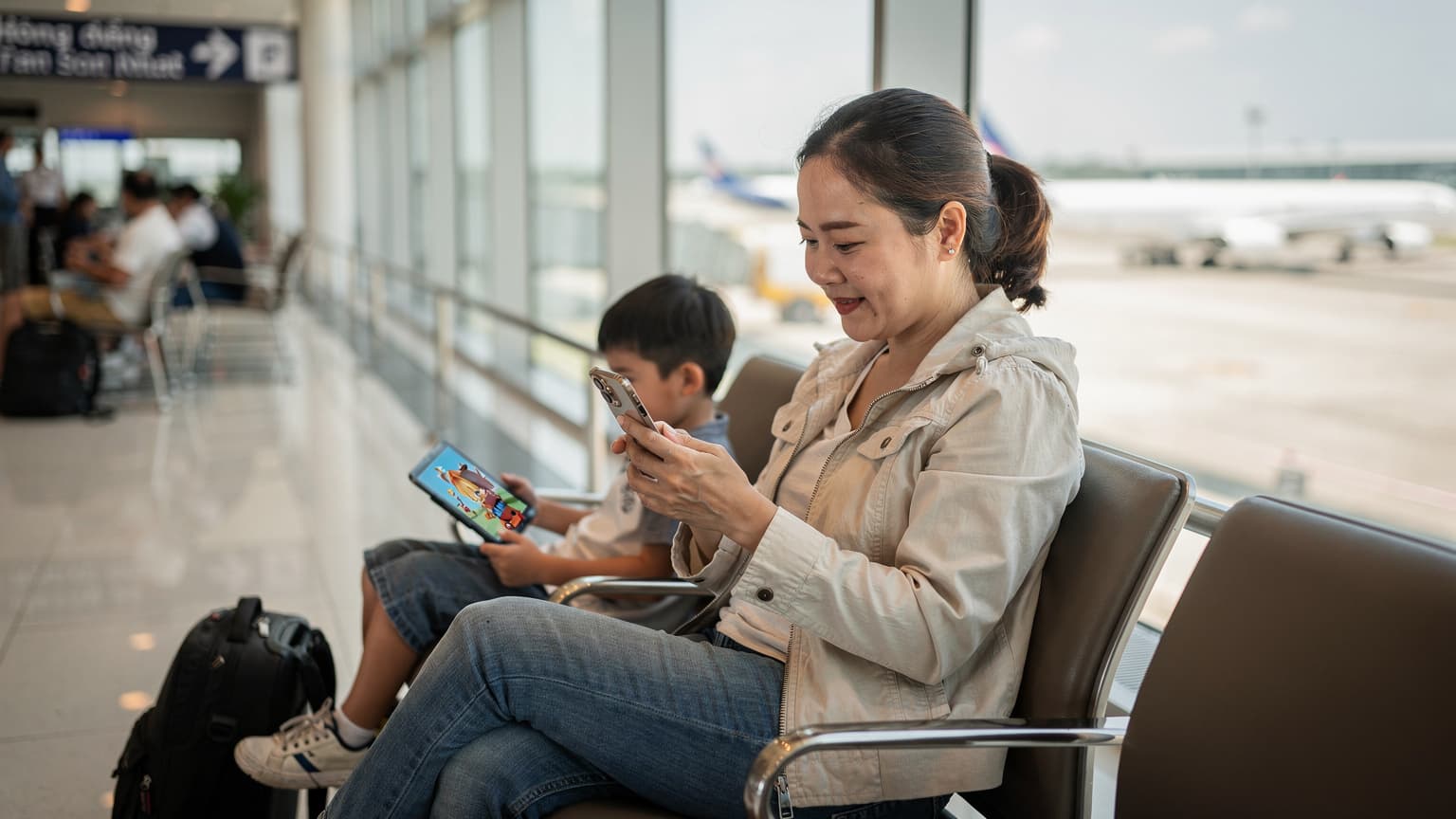 Một người phụ nữ Việt Nam đang ngồi tại khu vực chờ của sân bay Tân Sơn Nhất, chăm chú nhìn vào màn hình iPhone 11 của mình với biểu cảm nhẹ nhõm, bên cạnh là một vali và một chiếc iPad đang được một đứa trẻ sử dụng để xem phim hoạt hình.