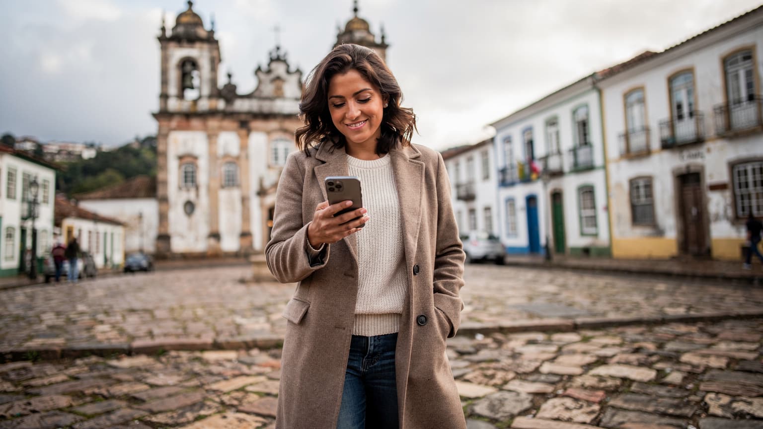 Uma mulher sorridente, vestindo um casaco de lã, segura seu smartphone enquanto caminha por uma rua de paralelepípedos em Ouro Preto, Minas Gerais, com as igrejas barrocas ao fundo sob um céu azul de inverno.