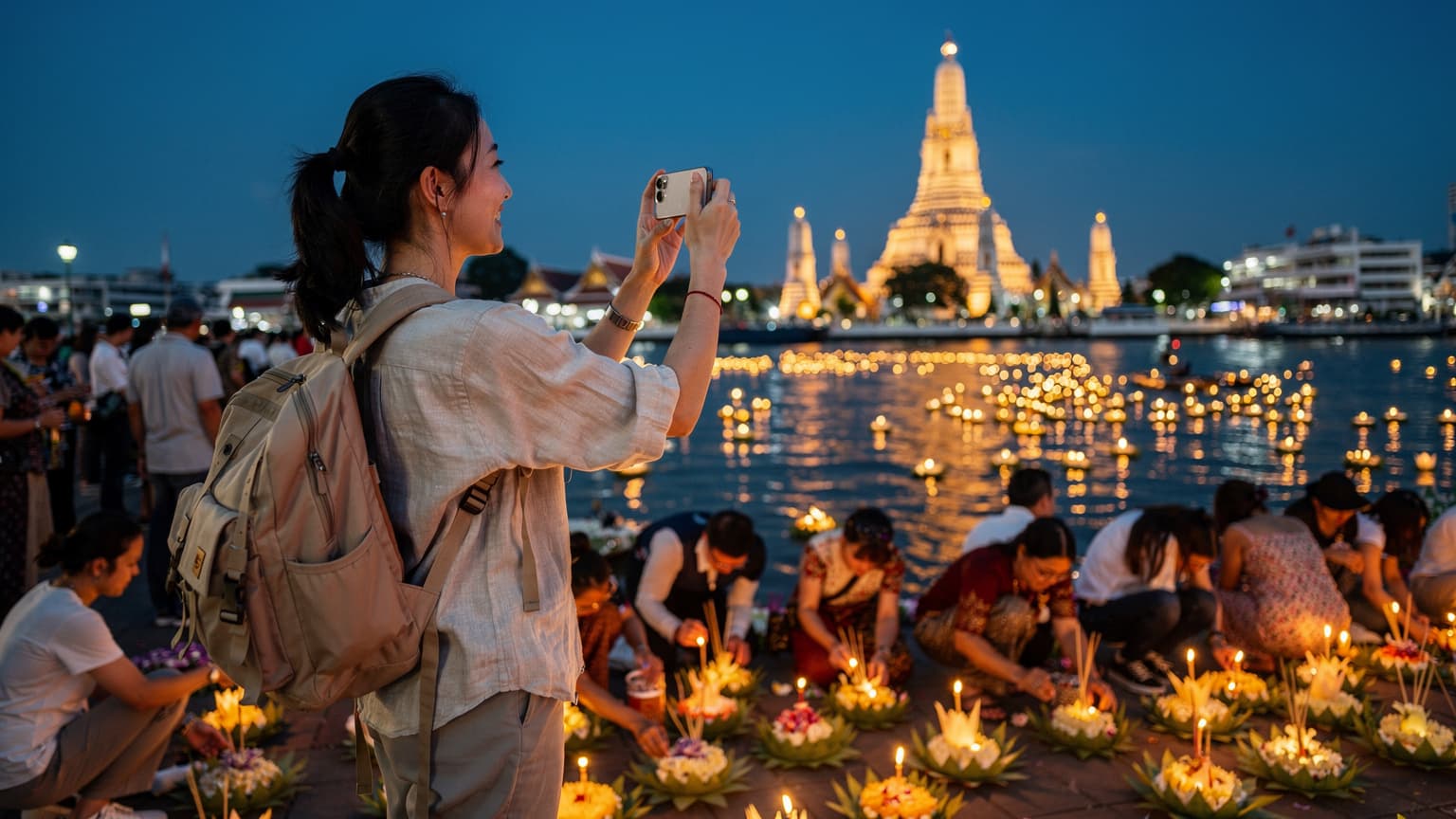 一位中国旅行者在曼谷水灯节期间,手持手机拍摄河灯,背景是寺庙灯火辉煌的夜景。