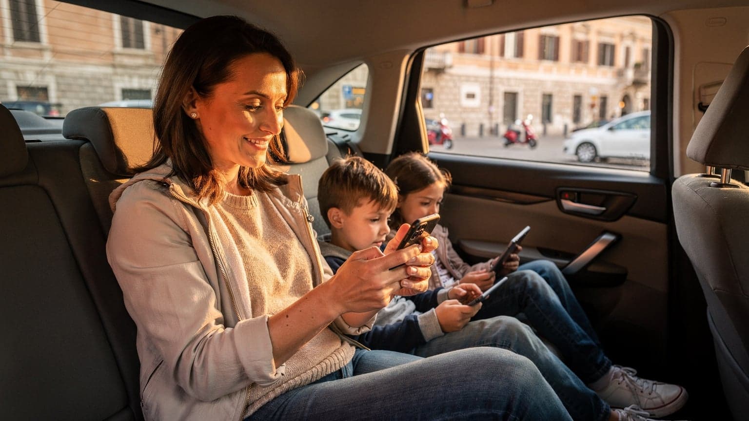 A mother calmly setting up her T-Mobile eSIM on her phone while her two children, ages 8 and 10, are engrossed in their tablets in the back seat of a taxi driving through a bustling European city street.