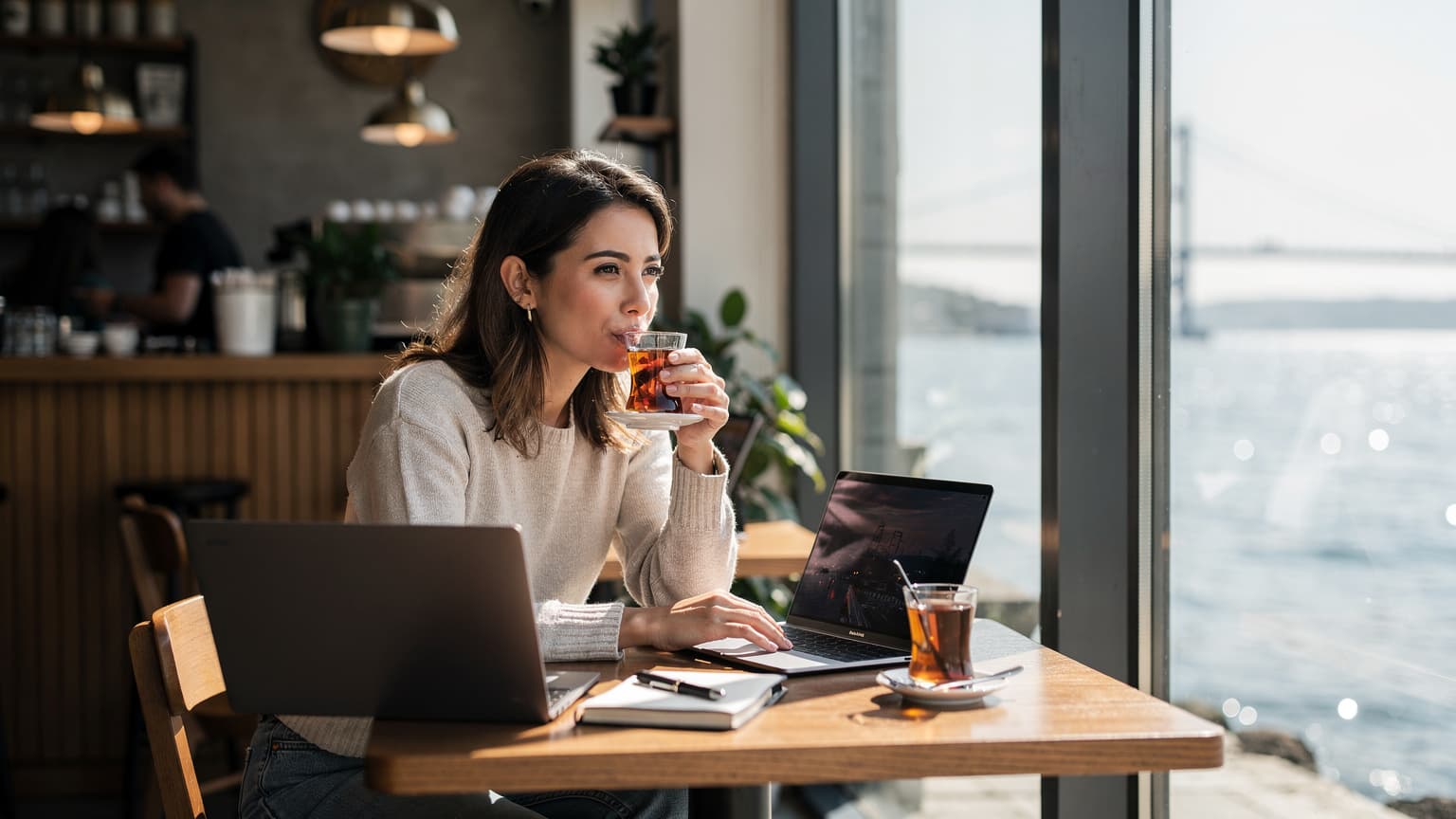 İstanbul'da bir kafede MacBook Air M3 ve Windows laptopları masada duran bir dijital göçebe kadın, pencereden Boğaz Köprüsü'ne bakarken kahvesini yudumluyor.