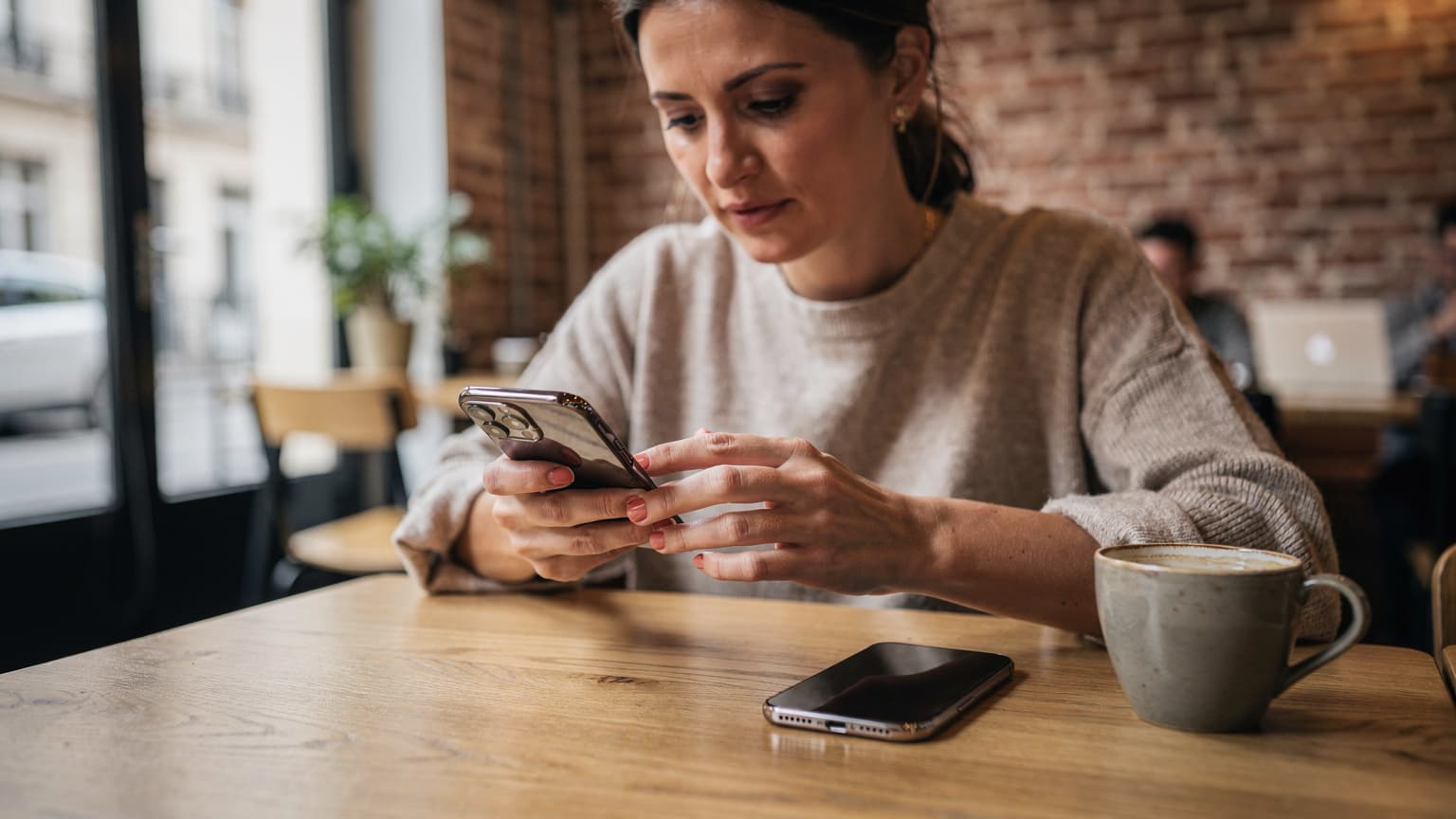 Une femme aux cheveux bruns, l'air concentré, transfère des données entre deux iPhones posés sur la table d'un café à la lumière du matin à Lyon, avec un café et un carnet à côté.