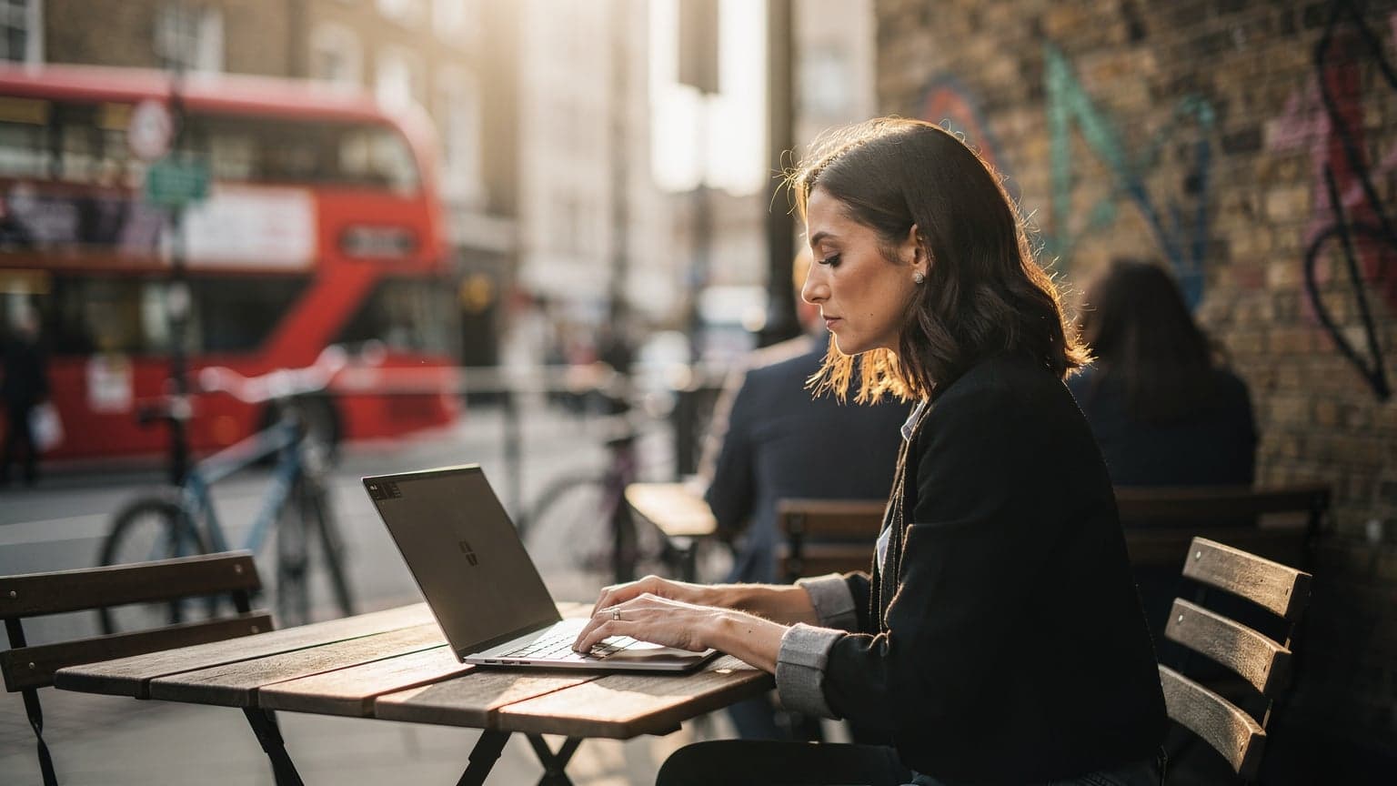 A woman working on her laptop at a sunlit outdoor cafe in London's Shoreditch, with a double-decker bus passing in the background.