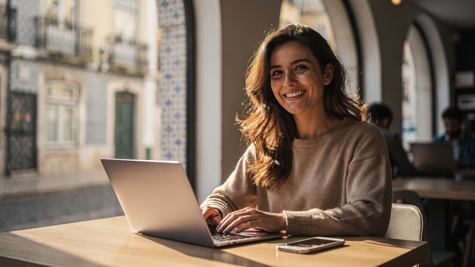 Uma mulher sorridente com um laptop e um smartphone, sentada num café moderno em Lisboa, com a luz dourada da manhã a entrar pelas janelas grandes, mostrando edifícios históricos ao fundo.