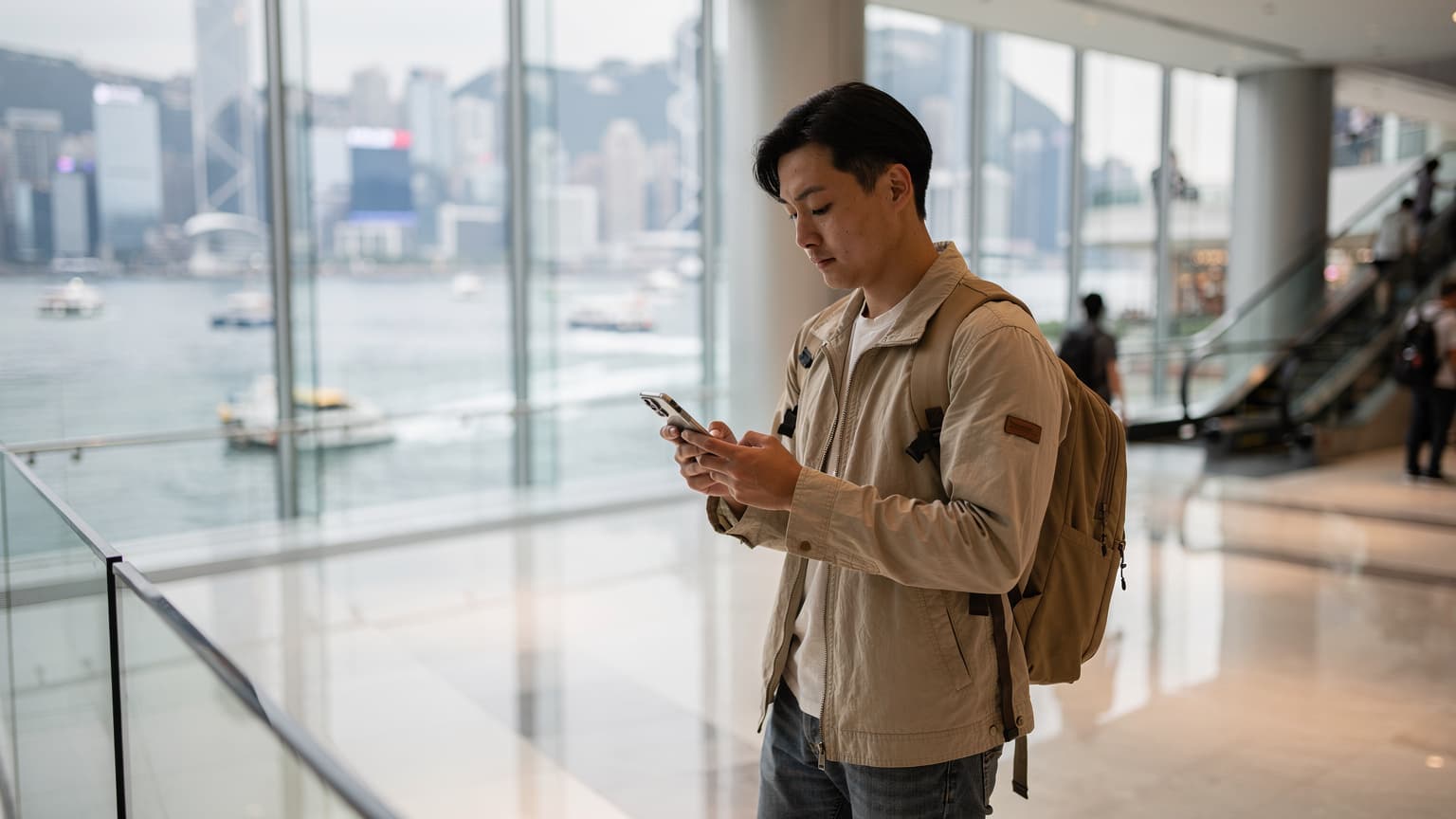一名中国大陆游客在香港中环IFC商场内使用智能手机查看eSIM信息,背景是维多利亚港的繁忙景象。