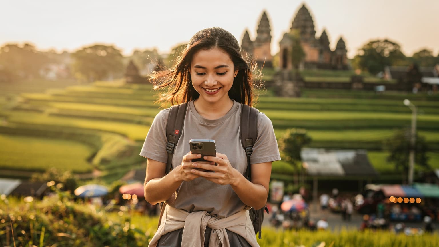 A young person, possibly Vietnamese, stands on a scenic overlook in a vibrant Southeast Asian landscape, holding a modern smartphone. They are looking at the screen with a relaxed, confident smile. The background, slightly out of focus, shows terraced rice fields or ancient temple ruins under natural golden hour light.