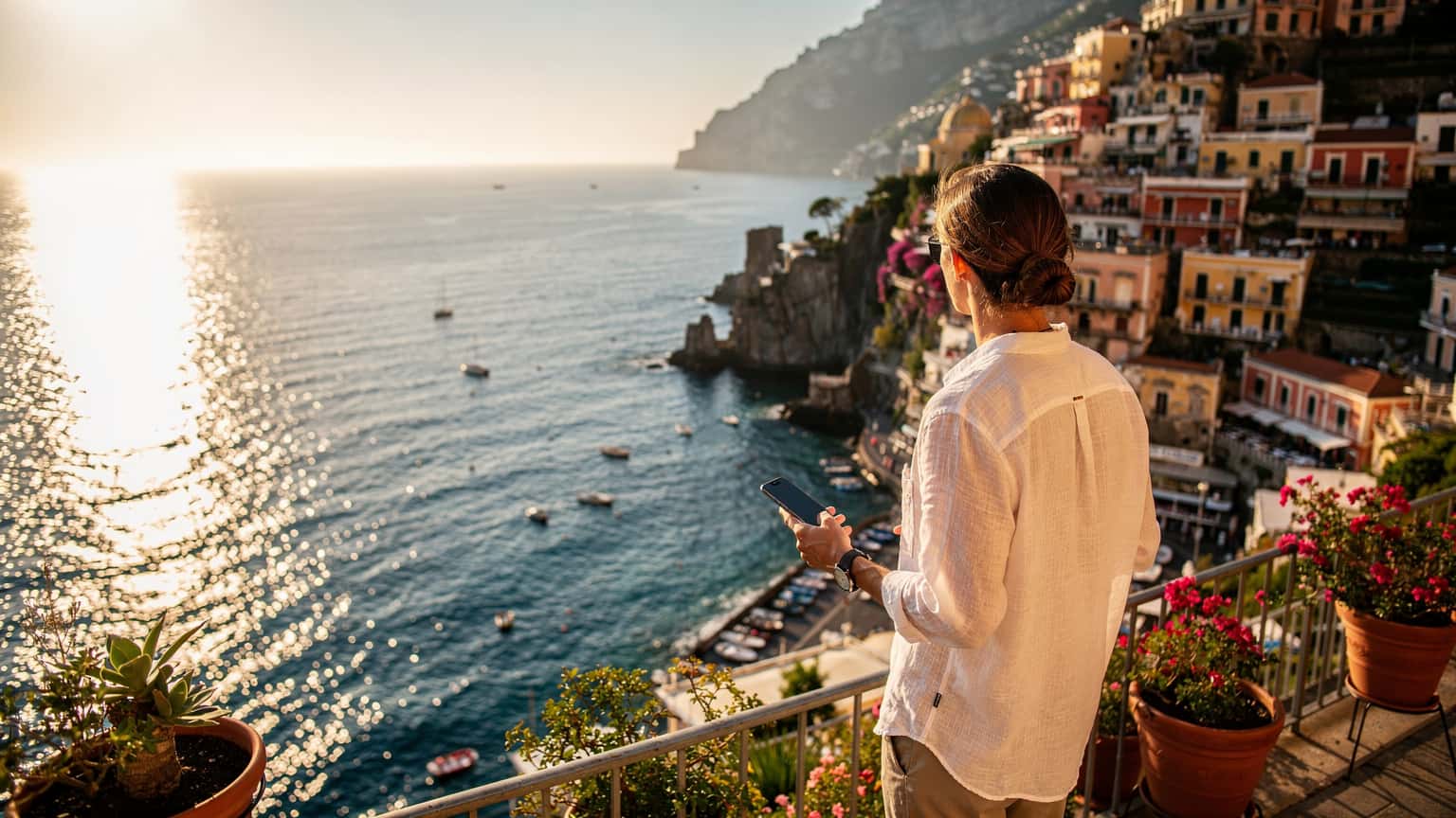 A person standing on a terrace overlooking Positano and the Amalfi Coast at sunset, holding a smartphone, with the Mediterranean Sea in the background.