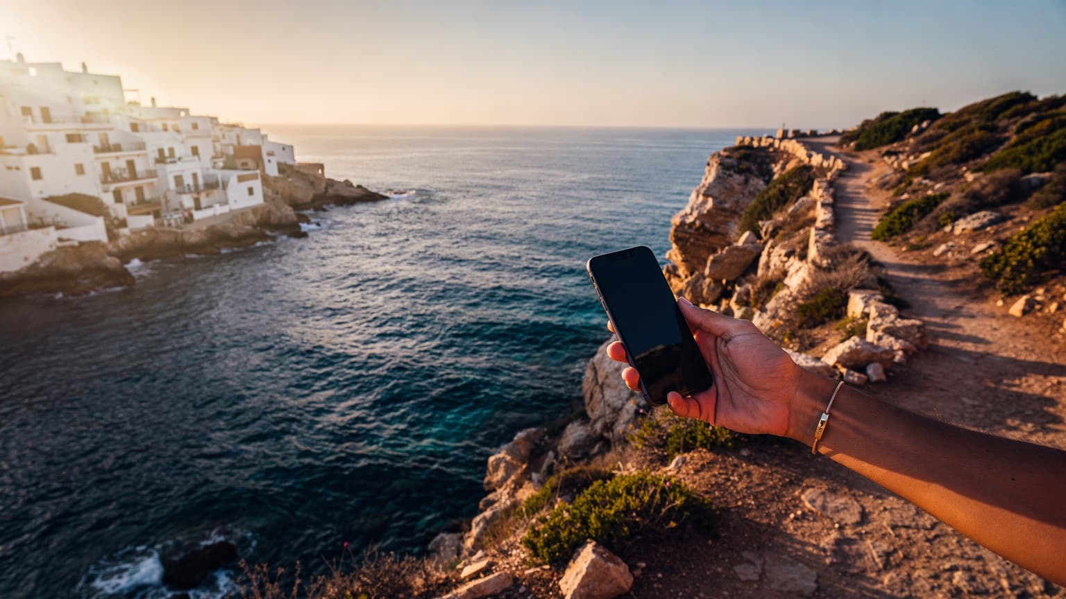 A photorealistic, editorial hero image of a hand holding a modern smartphone against a beautiful, sun-drenched Spanish coastal background, symbolizing effortless and connected travel.