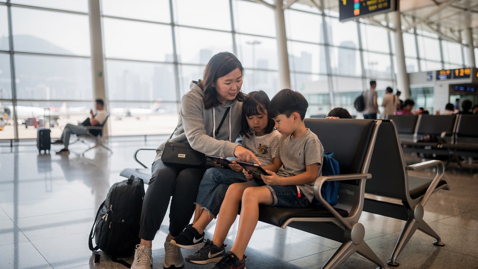 一位母親在香港機場的候機廳裡,正在幫助她的兩個孩子在 iPad 上設定旅行遊戲和離線內容,身邊放著登機箱和一個裝滿零食的背包。