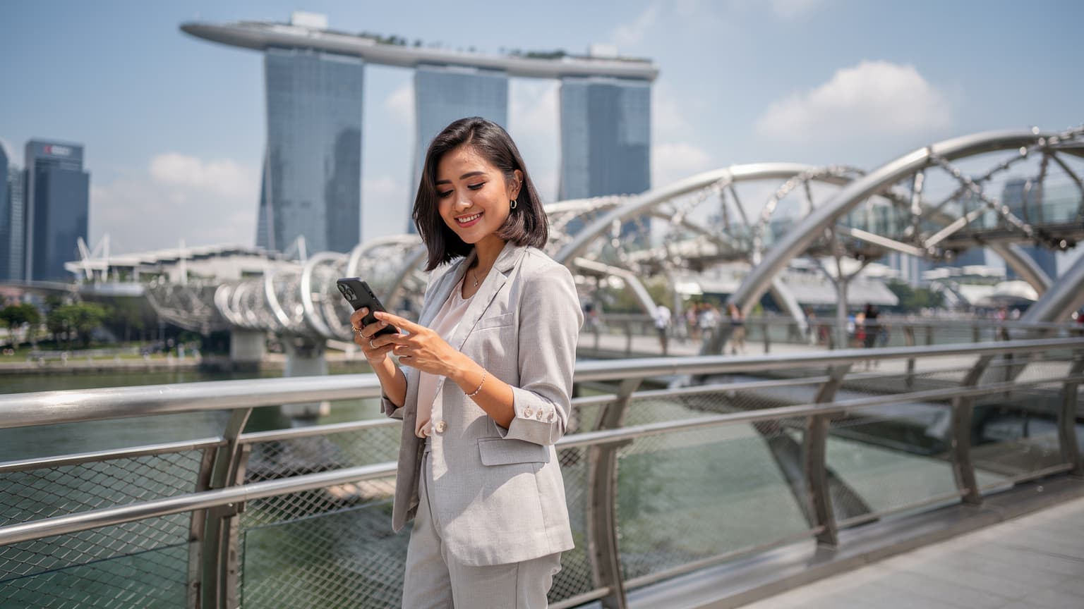 Seorang wanita muda Indonesia yang tersenyum sedang memegang ponsel pintar di tangan kanannya, melihat ke arah jembatan ikonik Helix Bridge di Marina Bay Sands, Singapura, dengan latar belakang langit biru cerah.