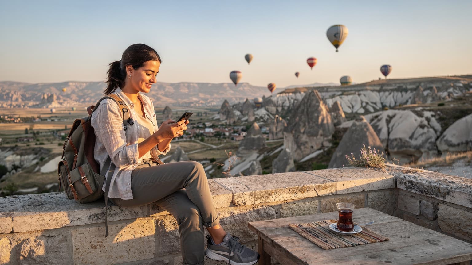 Un viajero revisa su teléfono mientras observa los globos aerostáticos al amanecer sobre los valles de Capadocia, Turquía, con una taza de té turco cerca.