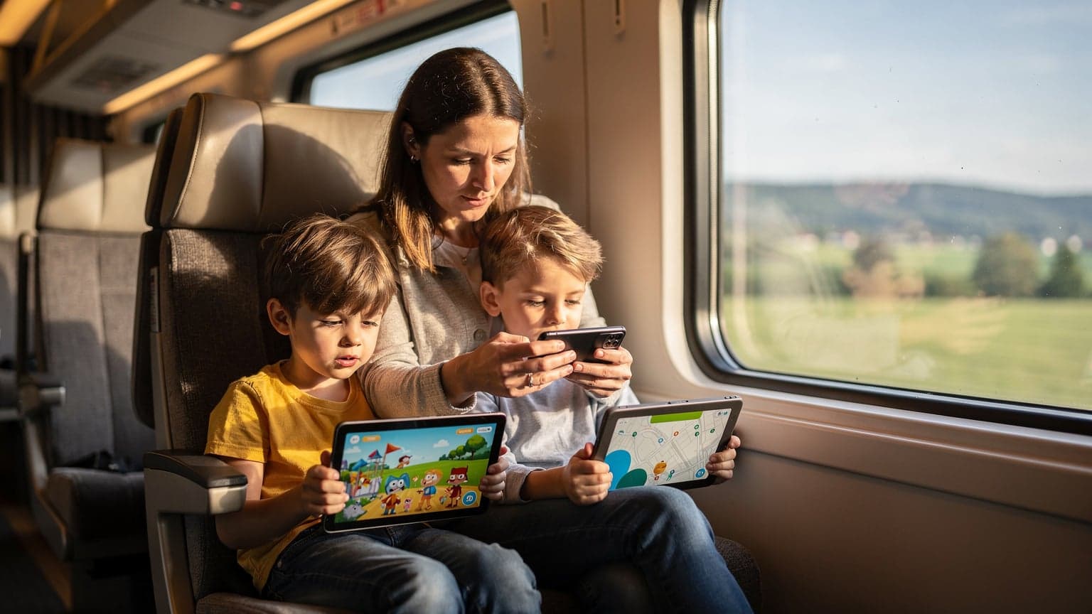Eine Mutter und ihre beiden Kinder im Alter von 8 und 11 Jahren sitzen gemütlich in einem Zugabteil der Deutschen Bahn, während die Landschaft vorbeizieht. Die Kinder schauen auf ihre Tablets, auf denen scheinbar Disney+ läuft, während die Mutter auf ihrem Smartphone etwas plant. Alle Geräte sind über eine stabile Internetverbindung verbunden.
