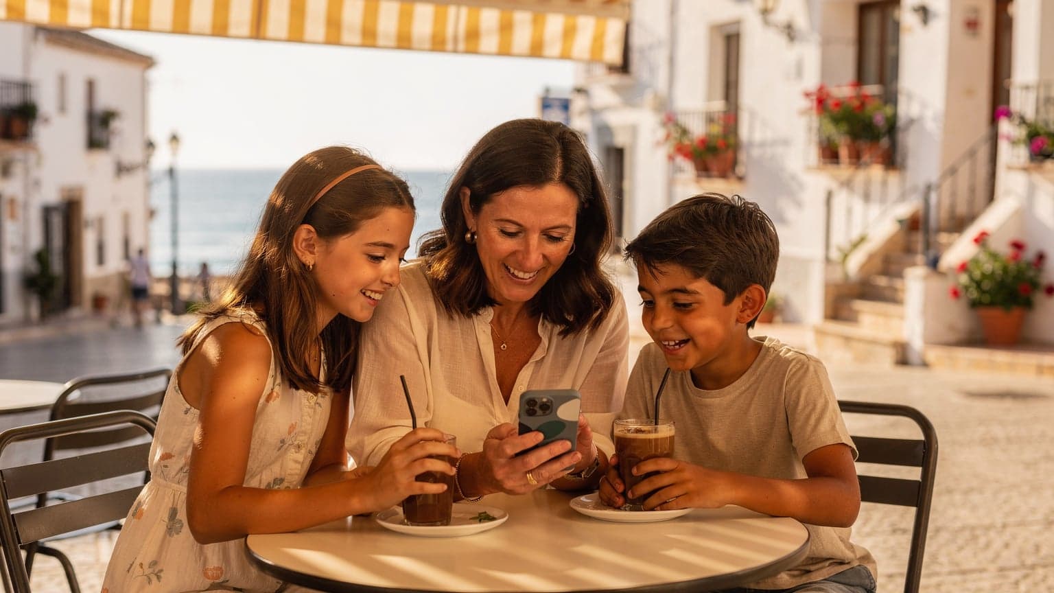 Una madre sonriente con sus dos hijos, un niño y una niña, sentados en un café al aire libre en un pueblo costero andaluz, mientras la madre les muestra algo divertido en su teléfono móvil con una eSIM activa.