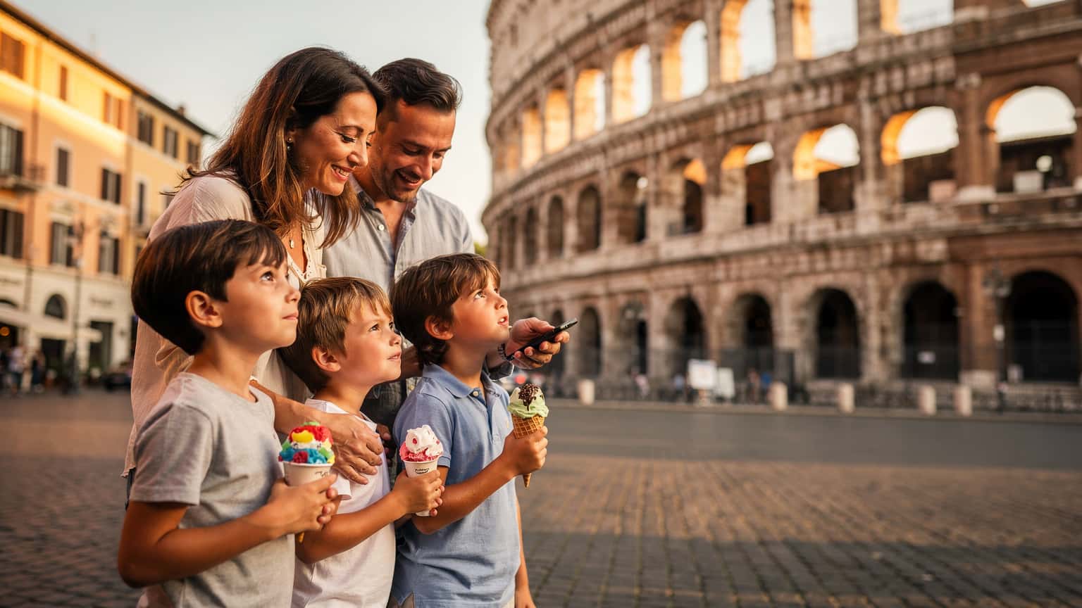 Une famille avec deux enfants, un garçon et une fille, souriante et détendue, admirant le Colisée de Rome au lever du soleil, tenant des glaces à l'italienne. Les parents vérifient discrètement leurs téléphones pour des cartes ou des informations.