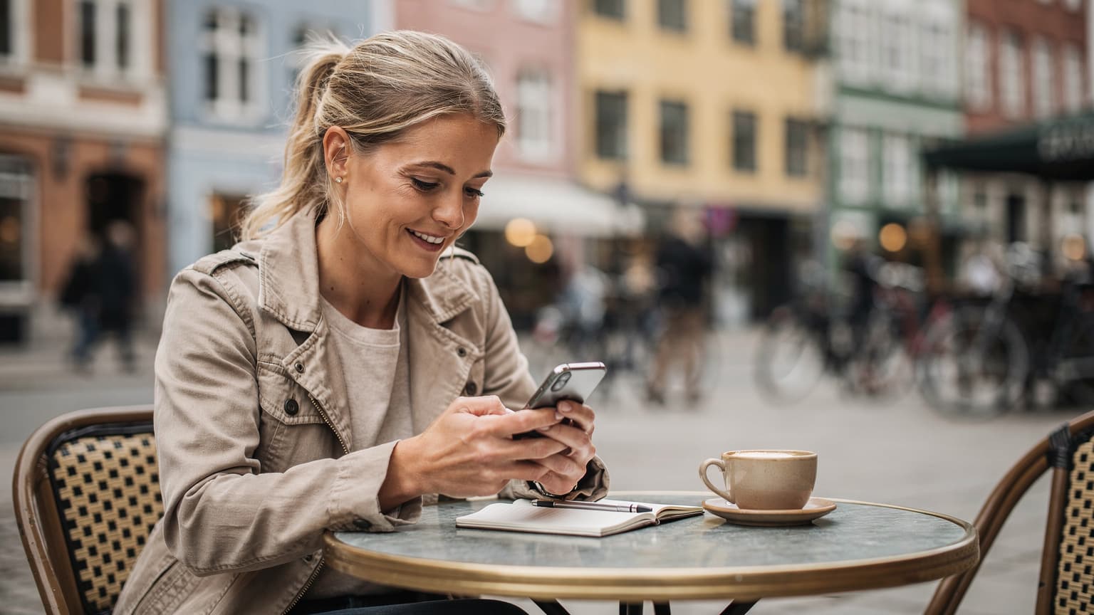En kvinde sidder på en travl gadecaf i København, kigger på sin iPhone med et smil, mens en kop kaffe og en notesbog ligger foran hende.