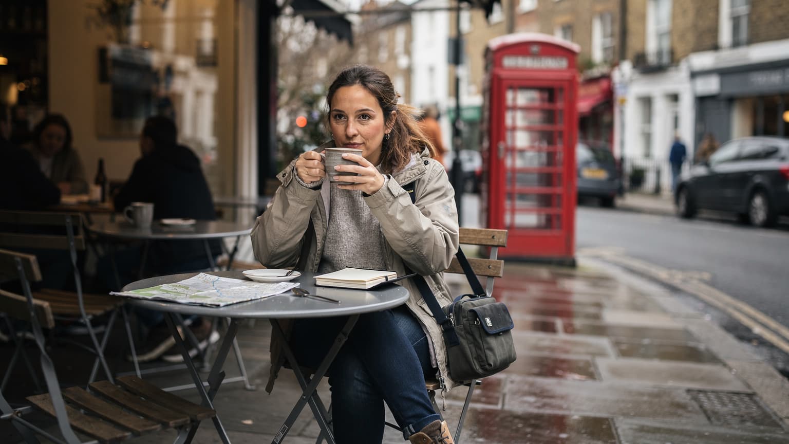 A woman sips tea at an outdoor cafe in London, with a classic red telephone box visible in the background on a slightly overcast afternoon.