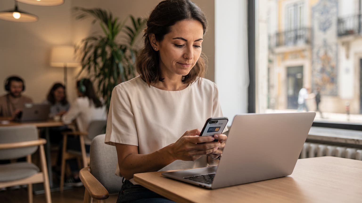 Une femme française, Antoine, vérifie son téléphone avec une carte eSIM sur l'écran dans un café de coworking animé à Lisbonne, les lumières de la ville visibles à travers une grande fenêtre, son passeport et une tasse de café à côté d'elle.